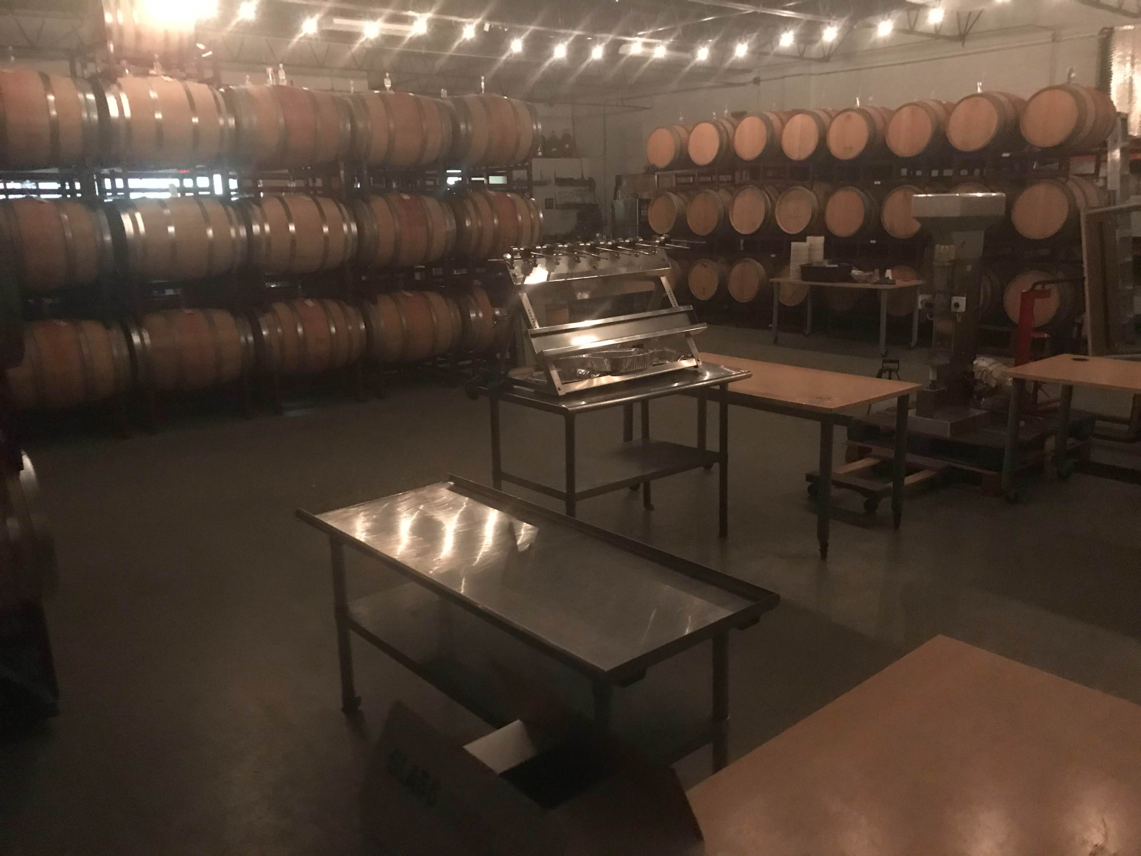 The image shows a dimly lit wine cellar with stacked wooden barrels along the walls and several stainless steel tables arranged in the center.