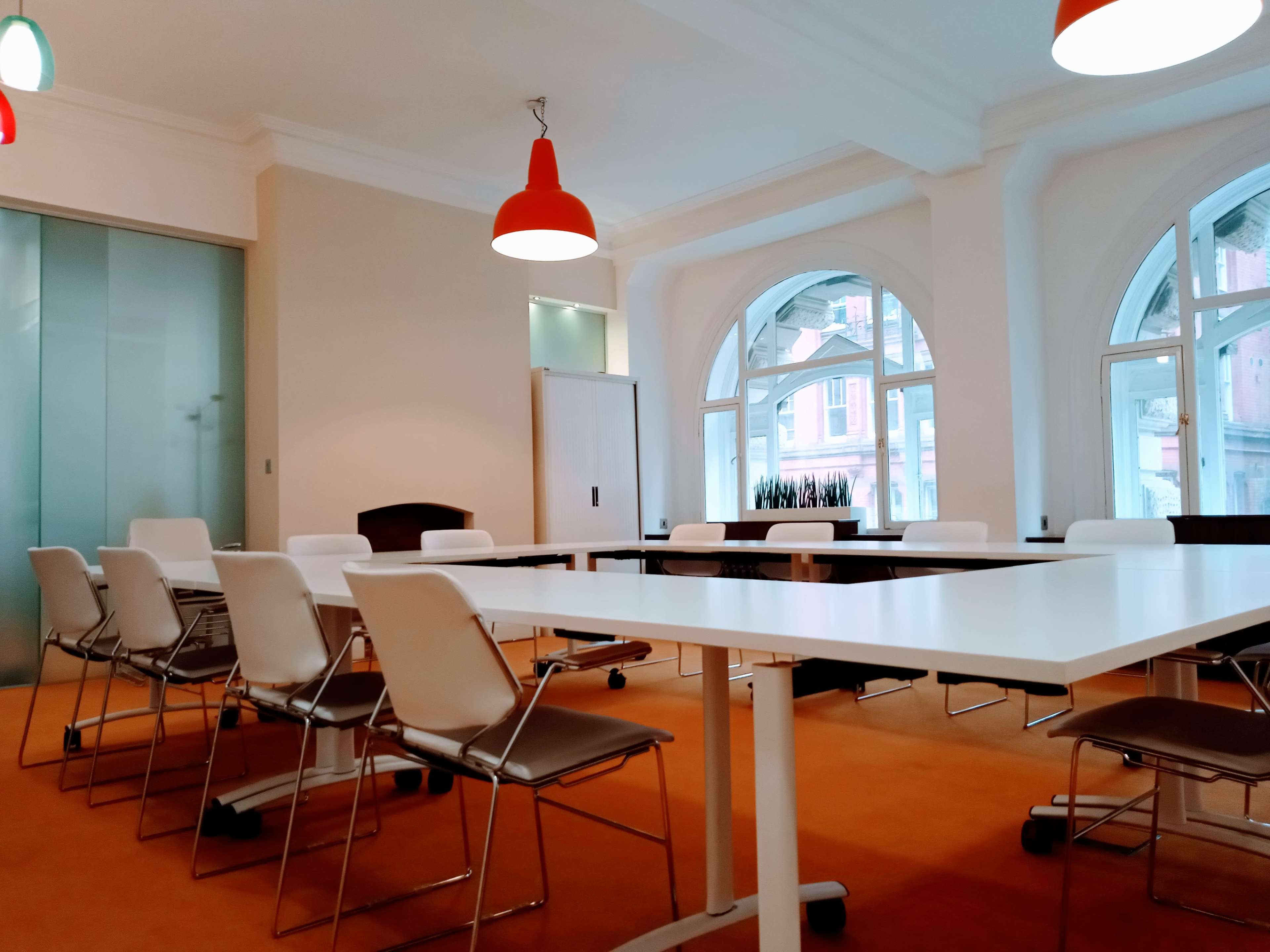 A modern conference room features a long table surrounded by chairs, large windows, and orange pendant lights.