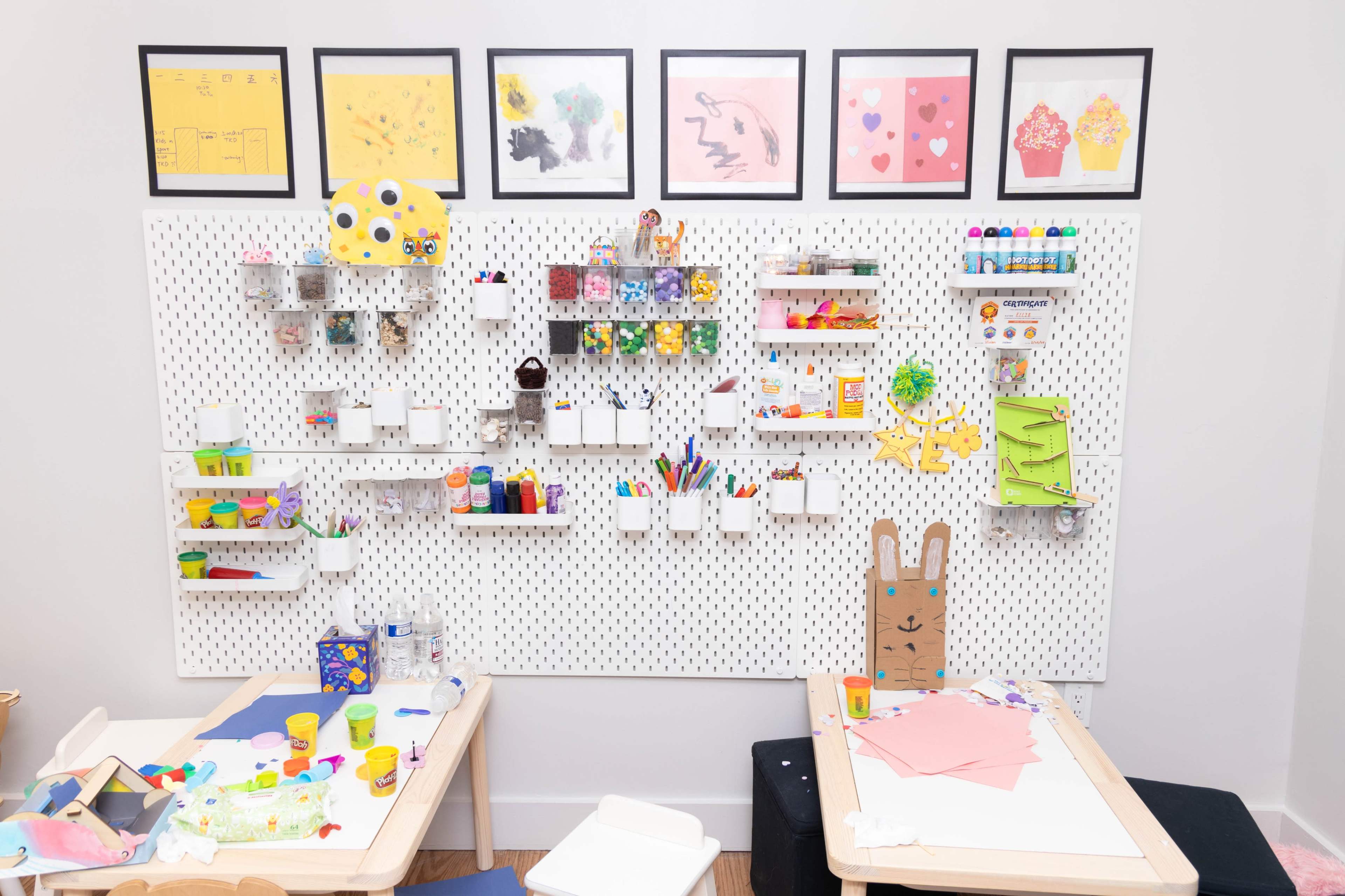 A craft room features a pegboard filled with art supplies organized in containers and colorful artwork displayed in frames.