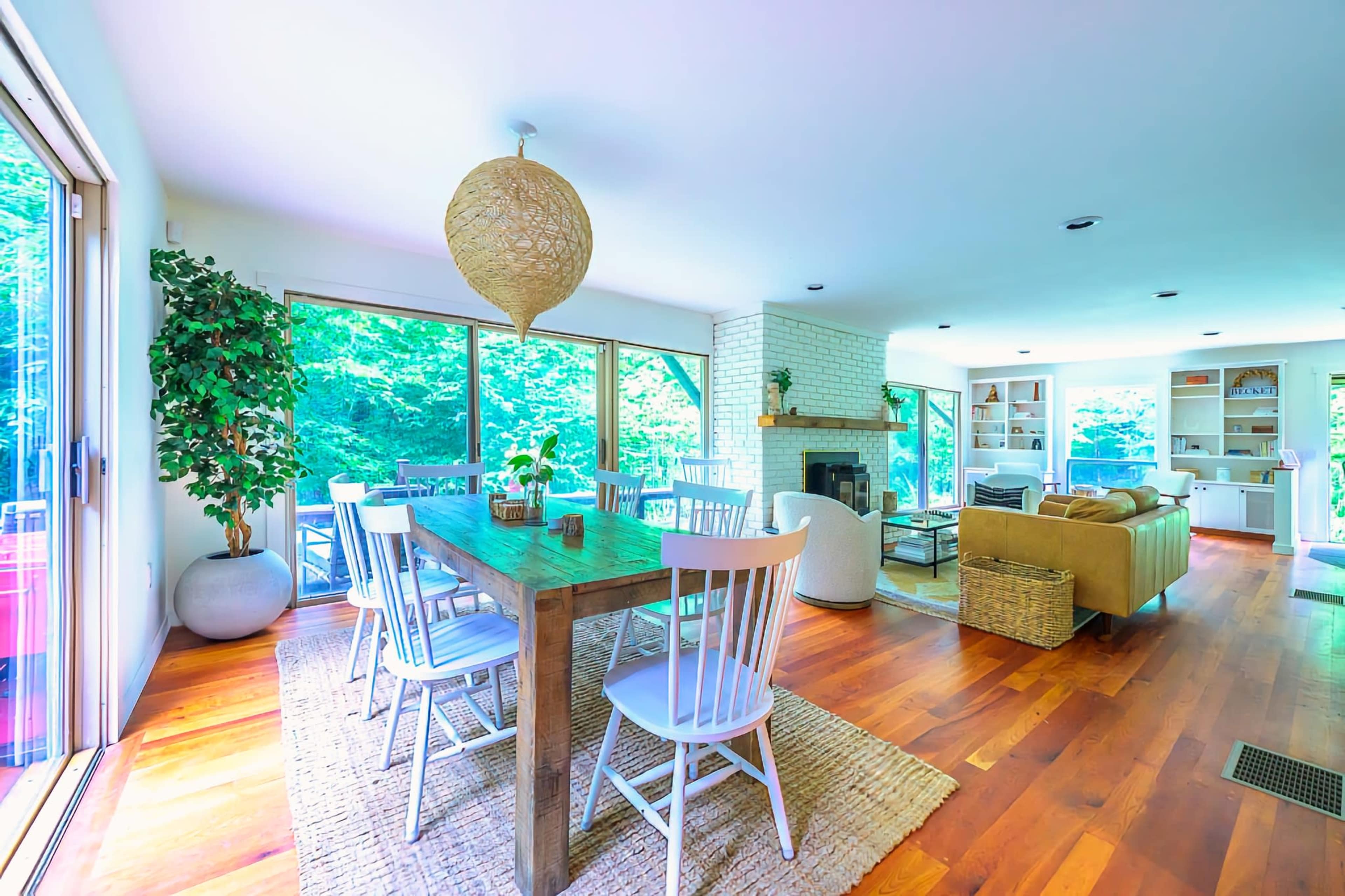 A bright, open living space featuring a dining table with white chairs, large windows overlooking greenery, and a cozy seating area with a sofa and fireplace.