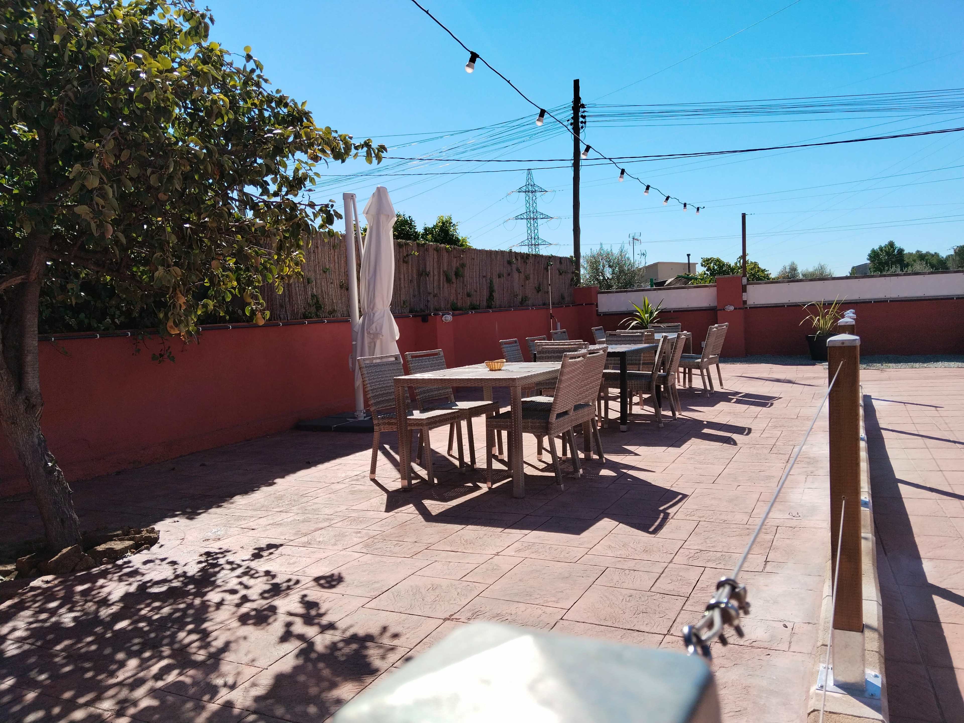 The image shows a sunny outdoor patio area with several wooden dining tables and chairs arranged beneath string lights and a large umbrella.