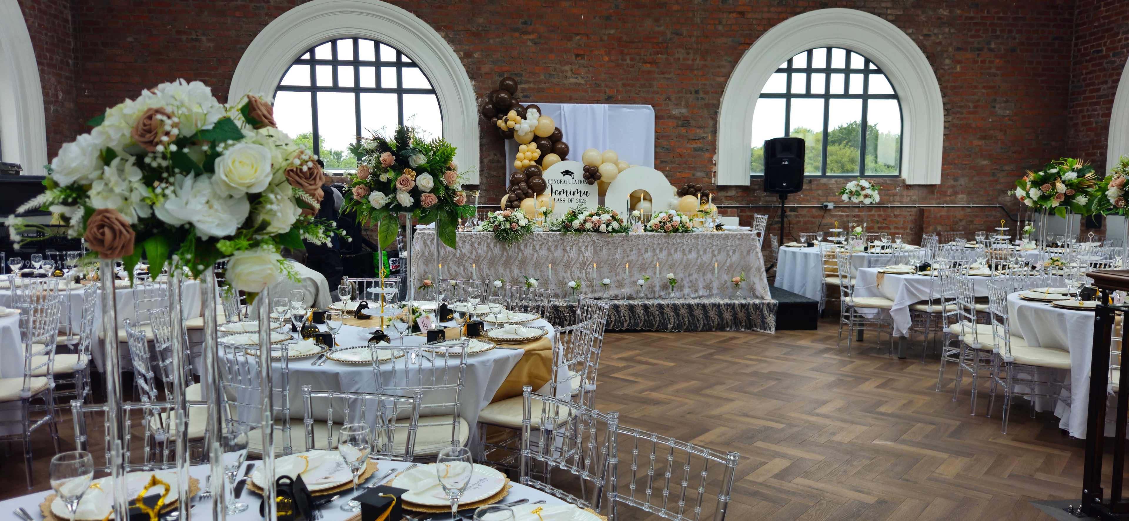 The image shows an elegantly decorated event space with round tables set for a celebration, featuring floral arrangements and a large elevated display at the front.