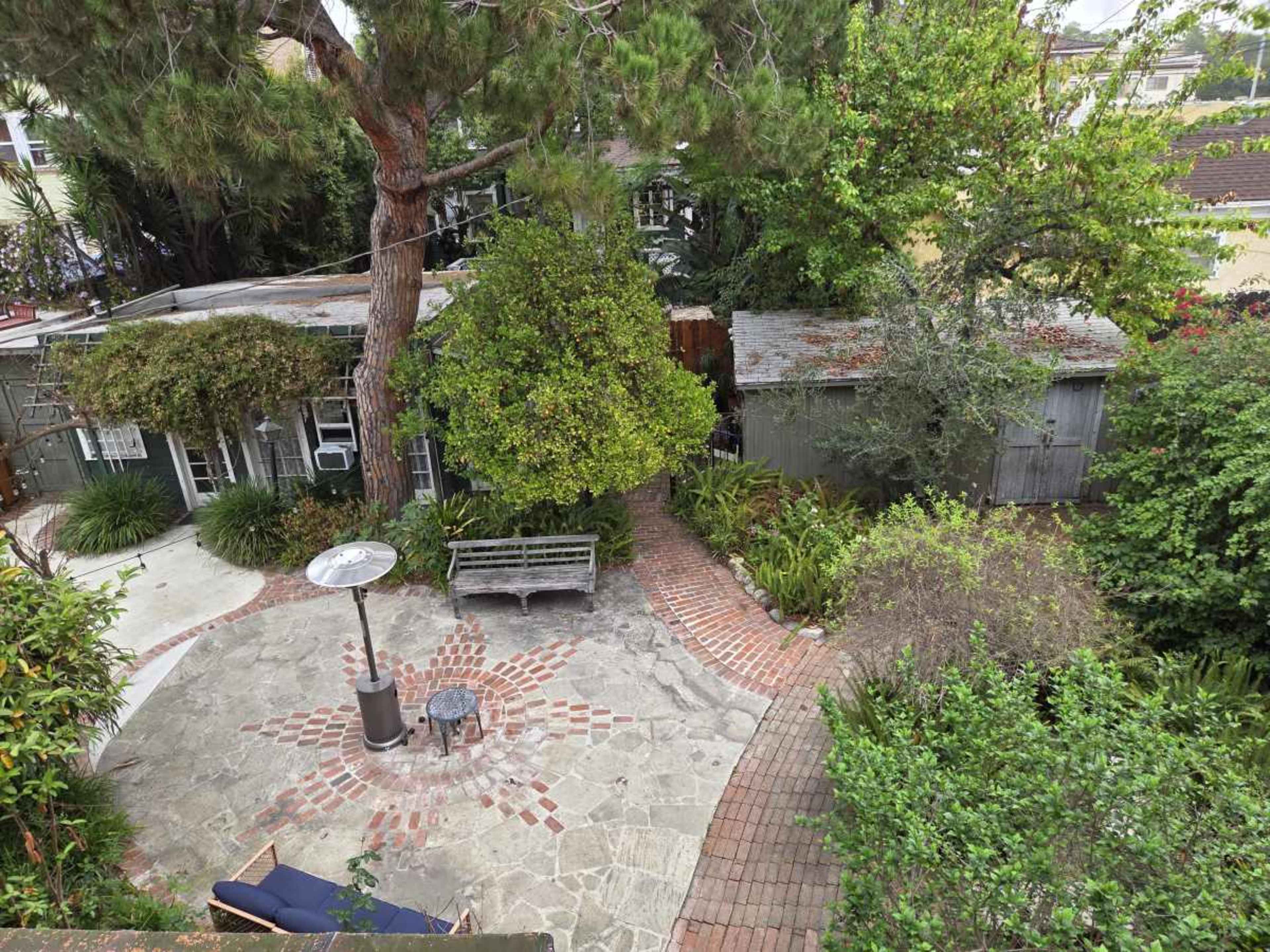 The image shows a landscaped backyard with a stone patio, a bench, a pathway, and a shed surrounded by trees and vegetation.