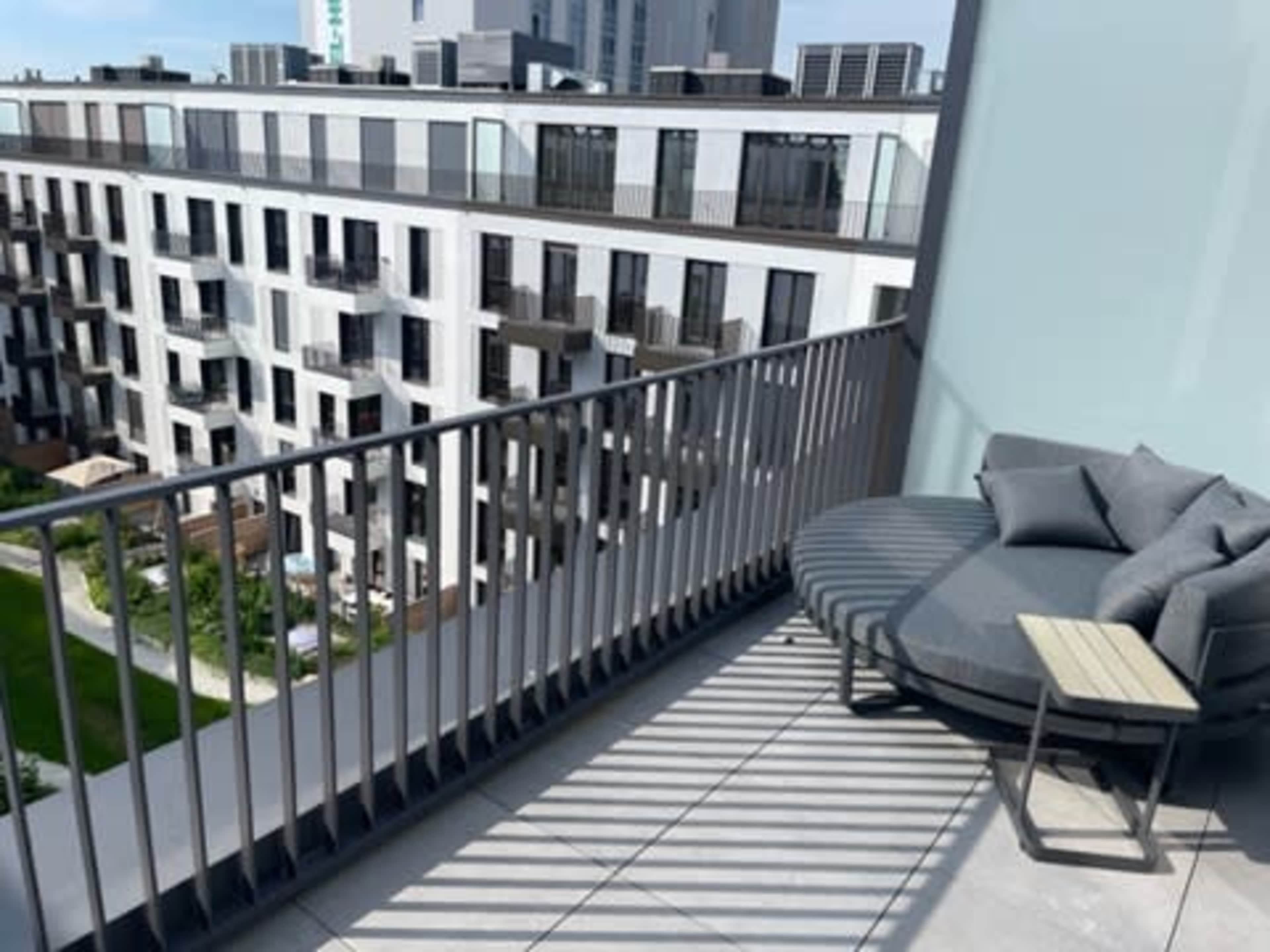 A modern balcony features a circular sofa and a small table, overlooking a multi-story building with a green courtyard below.