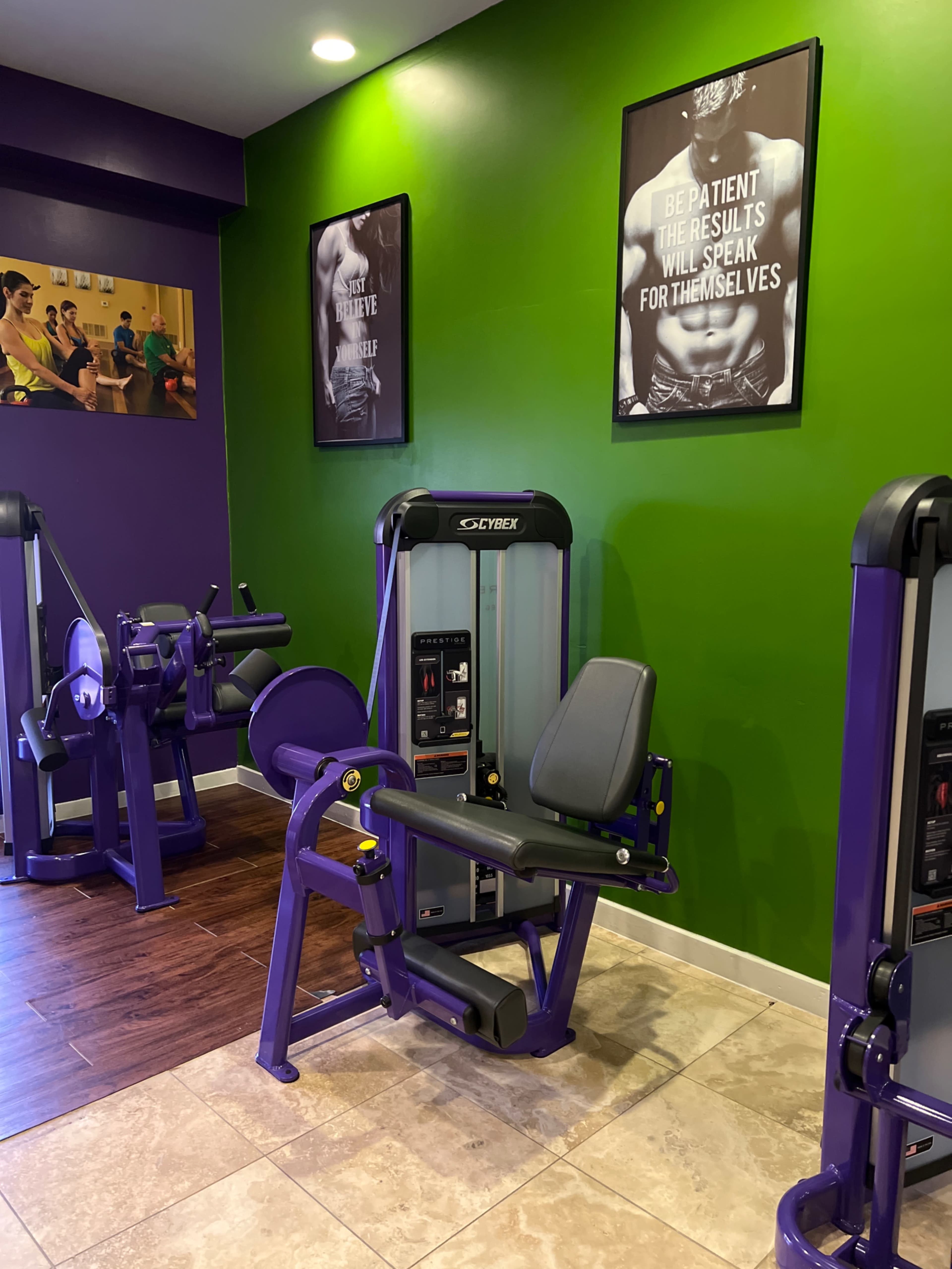 The image shows a gym interior with exercise equipment, including a leg press machine, set against a green and purple wall adorned with motivational posters.