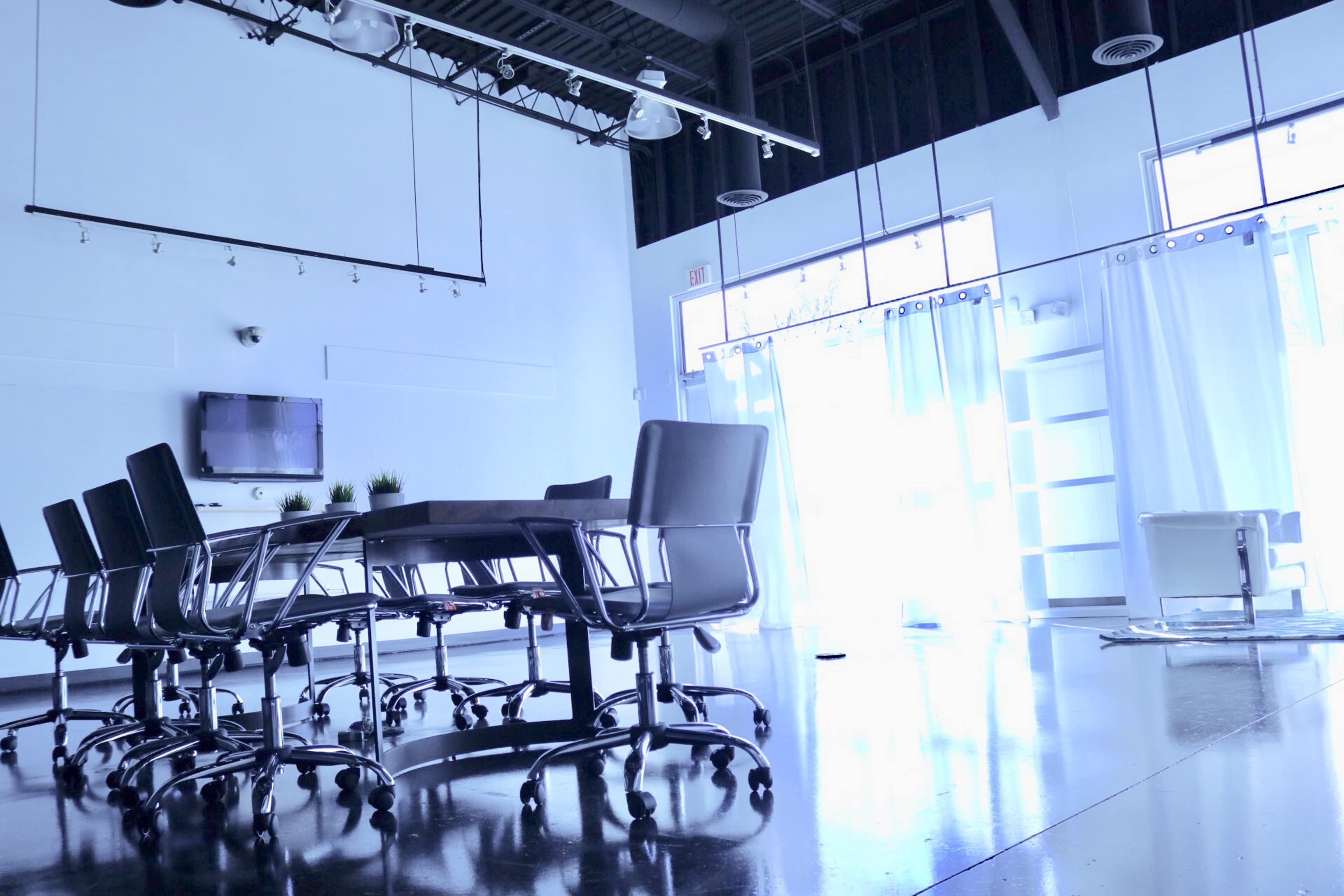 A modern conference room features a large table surrounded by black rolling chairs, with large windows and white curtains on one side.