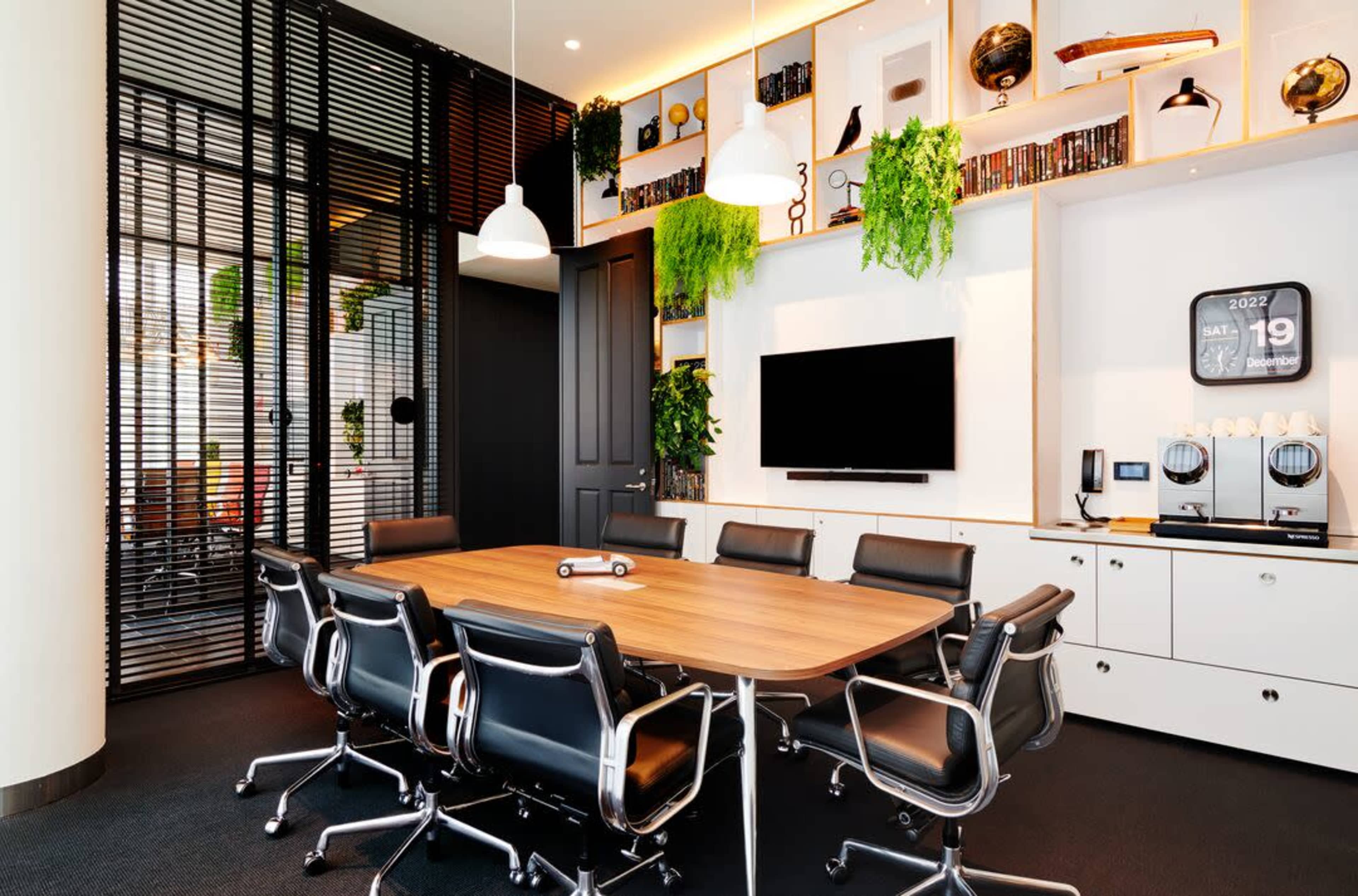 A modern conference room features a large wooden table surrounded by black chairs, with greenery on the walls and a television mounted across from the table.