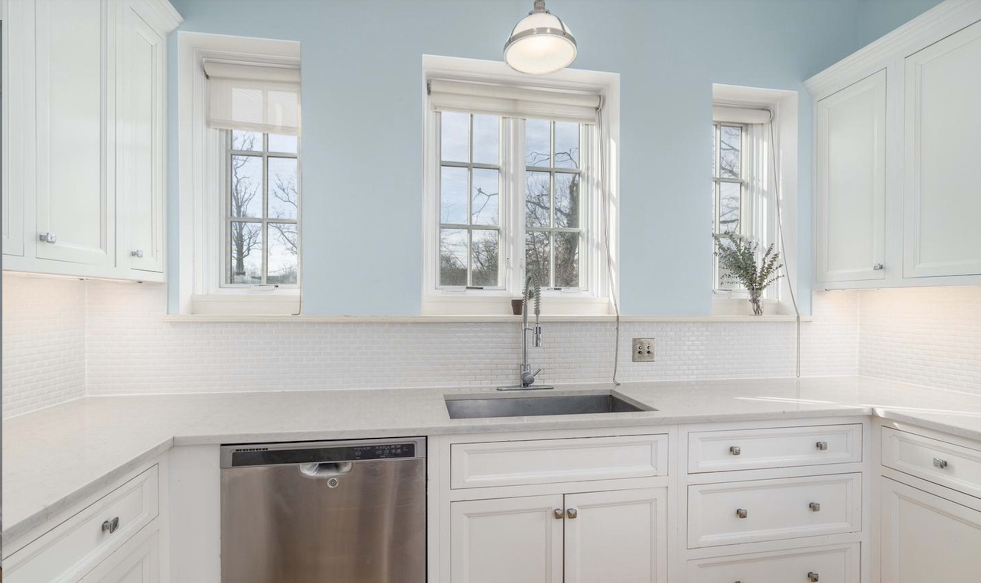 A clean kitchen features white cabinetry, a stainless steel dishwasher, and a single basin sink with windows above that provide natural light.