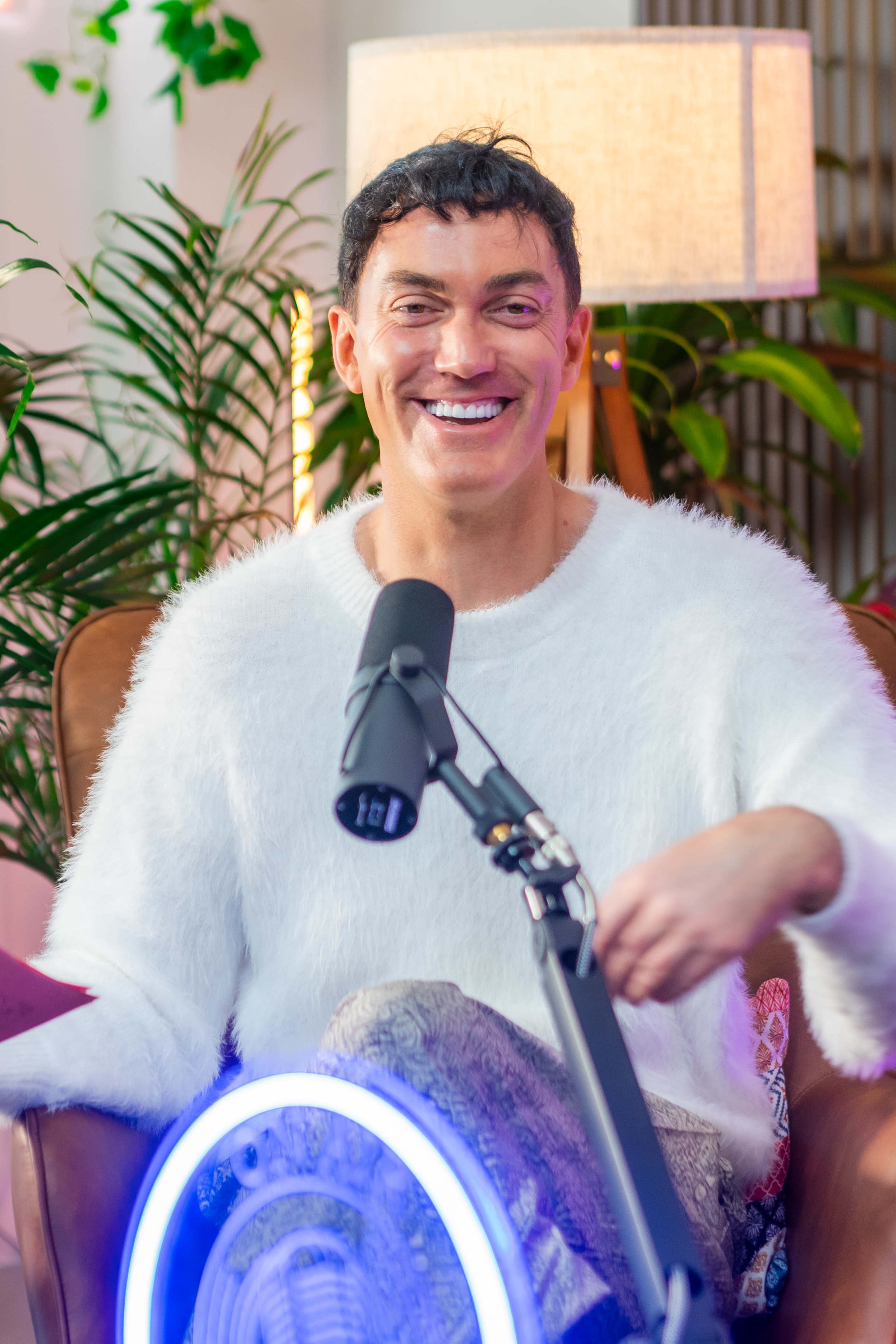 A person wearing a fluffy white sweater smiles while sitting in a chair, with a microphone and lamp visible in the background.