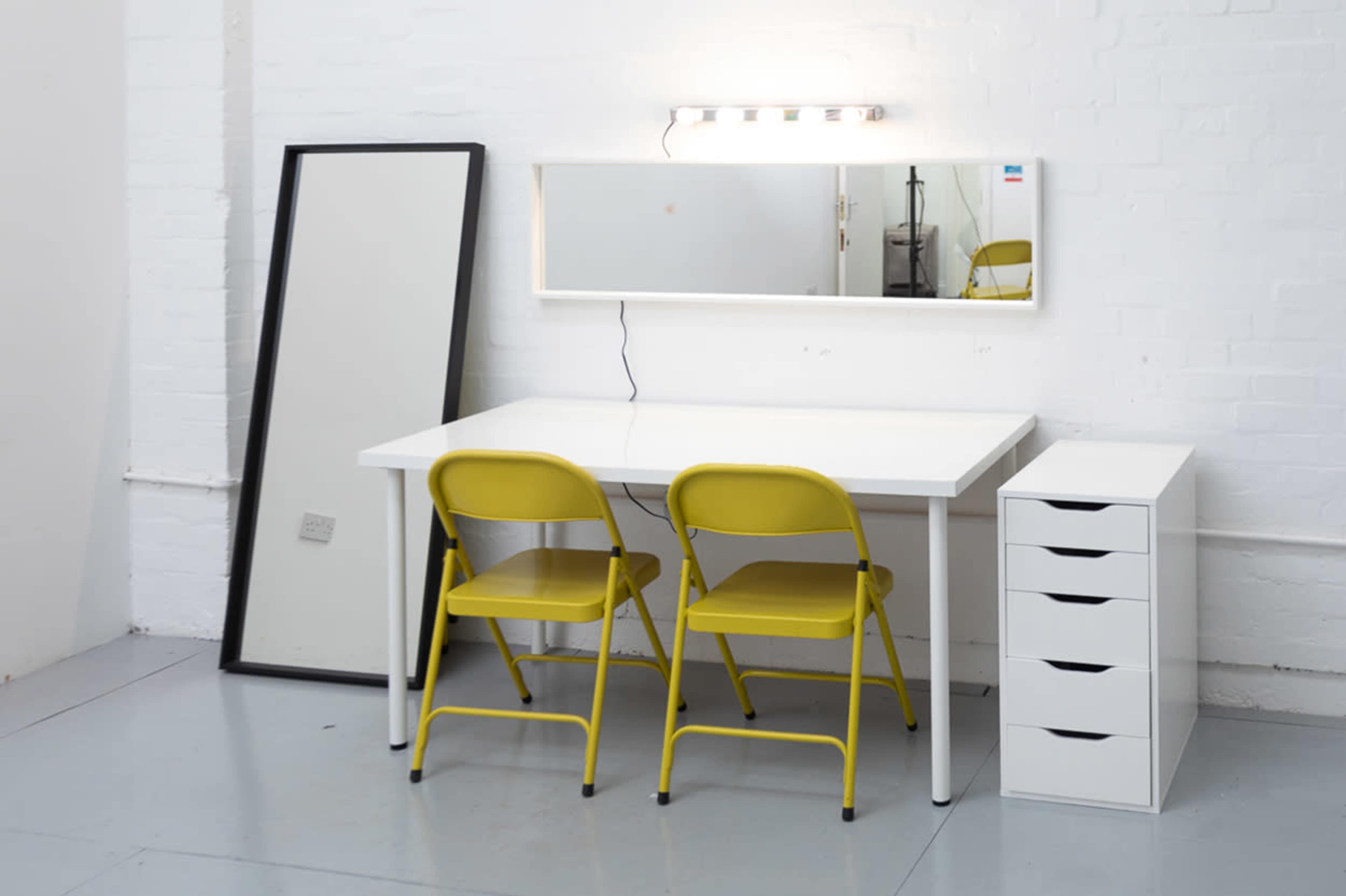 A white desk with two yellow chairs sits in front of a mirrored wall and a tall white storage unit in a bright room.