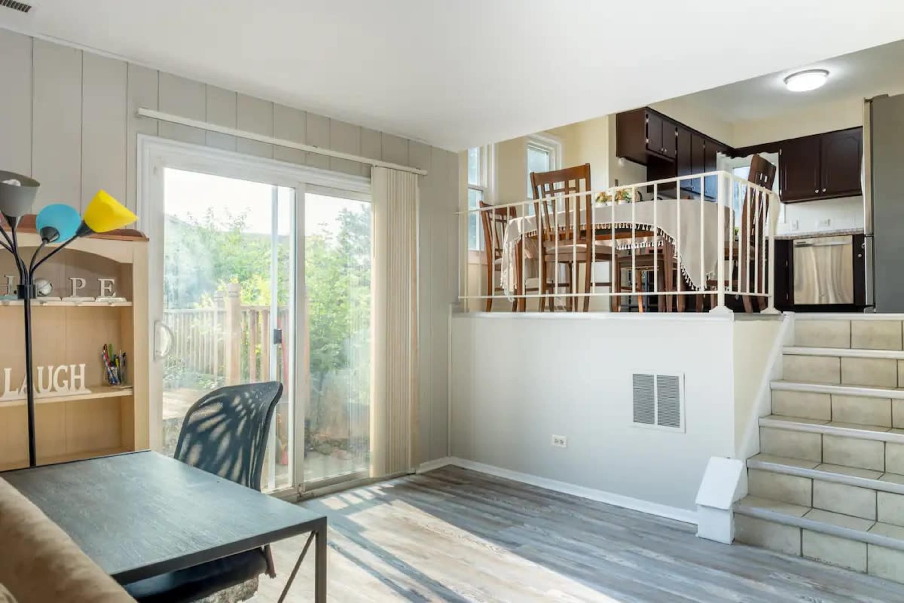 A bright living area features a sliding glass door leading to an outdoor space, with a dining area and kitchen visible on a raised platform.
