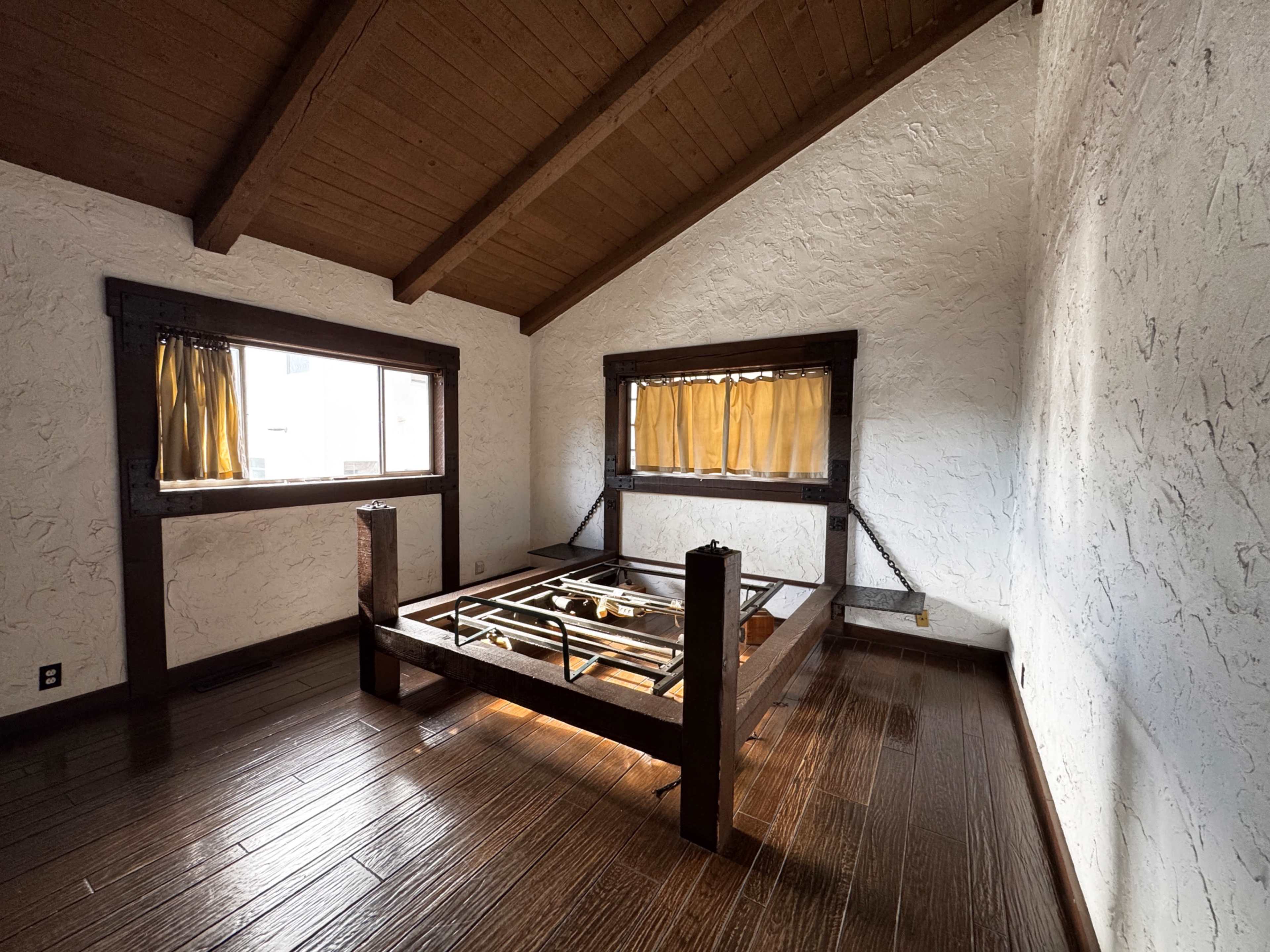 The image shows a sparsely furnished room with a wooden bed frame and two windows adorned with yellow curtains.