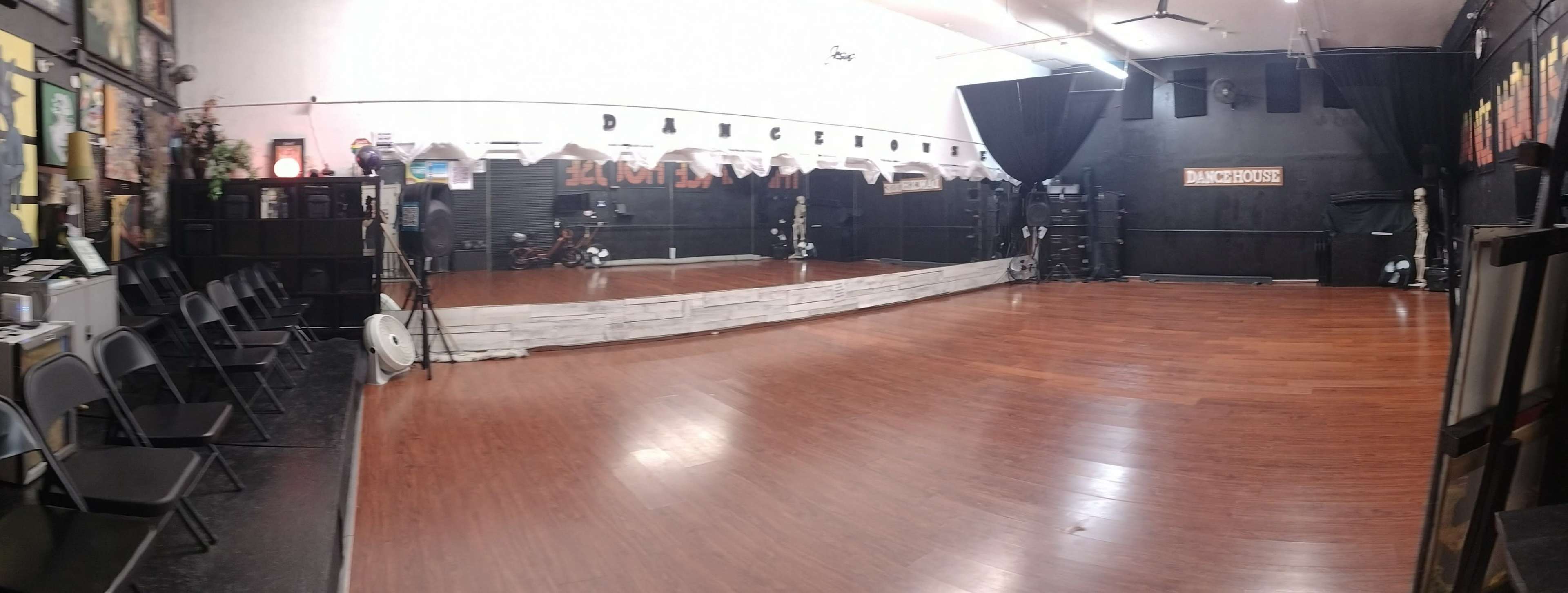 A spacious dance studio featuring a polished wooden floor, floor-to-ceiling mirrors along one wall, and chairs arranged along the side.