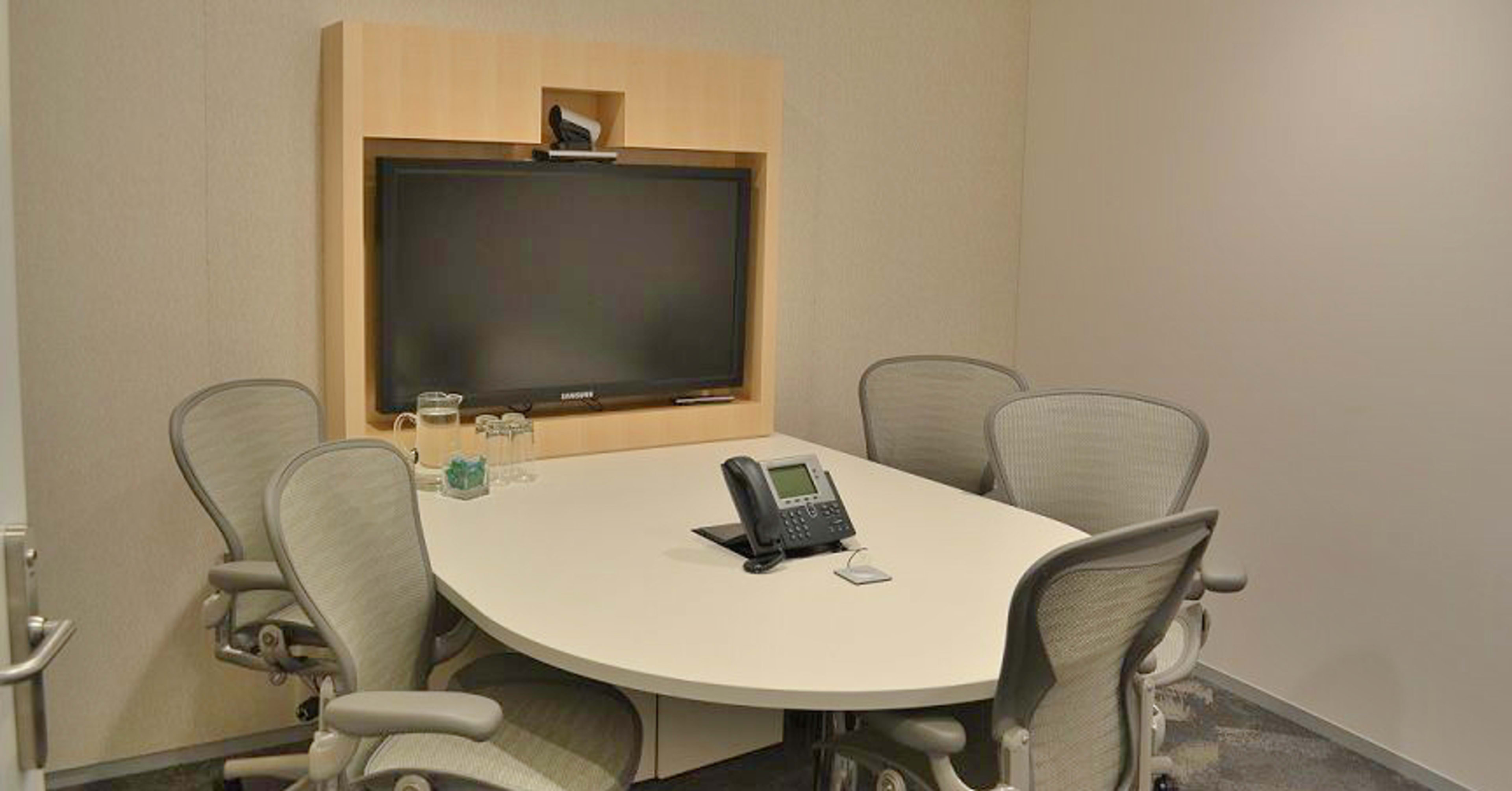 A small conference room with a large wall-mounted television, a round table, and four ergonomic chairs.