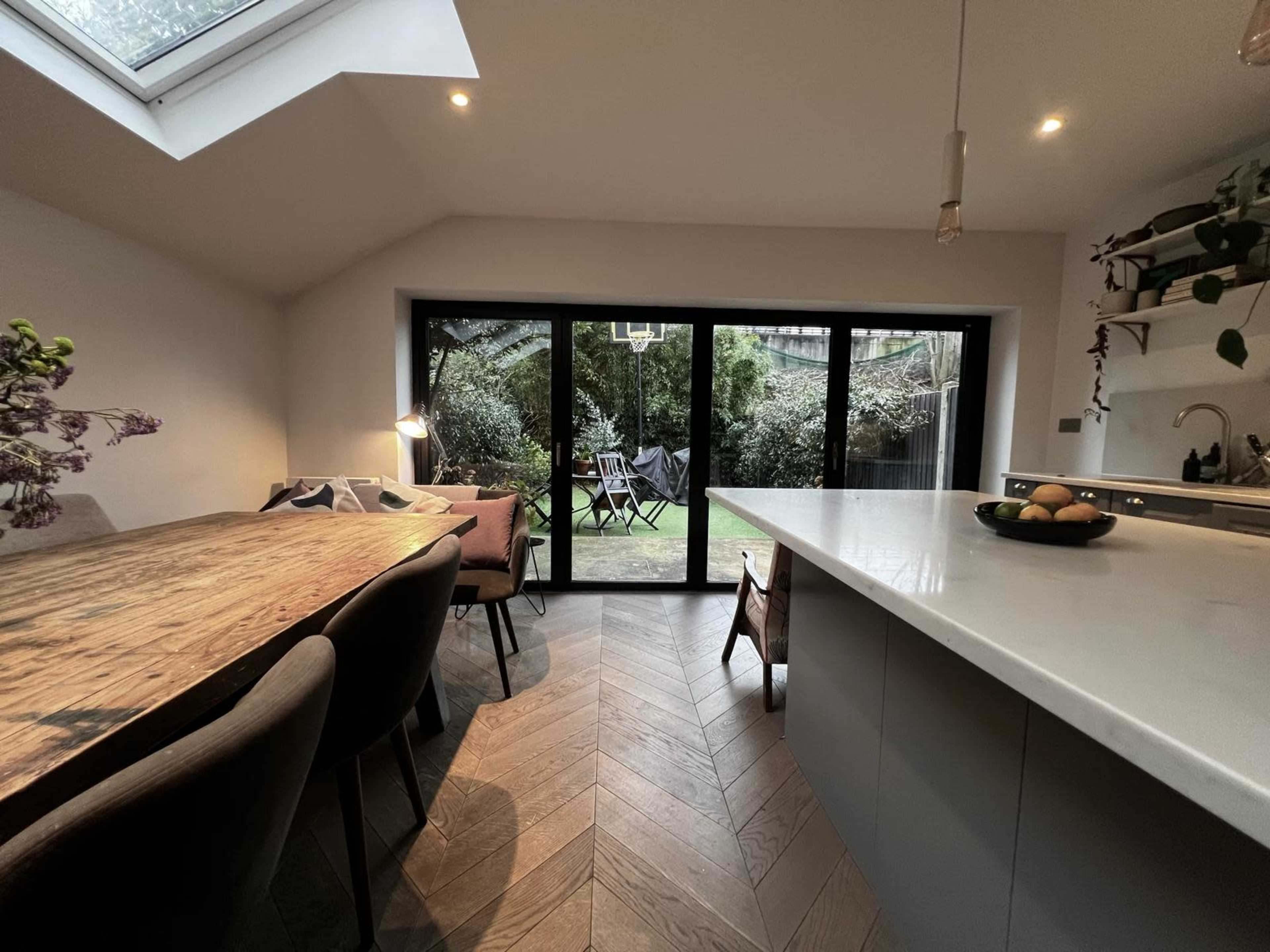 A dining area with a wooden table and chairs opens to a garden through large glass doors.
