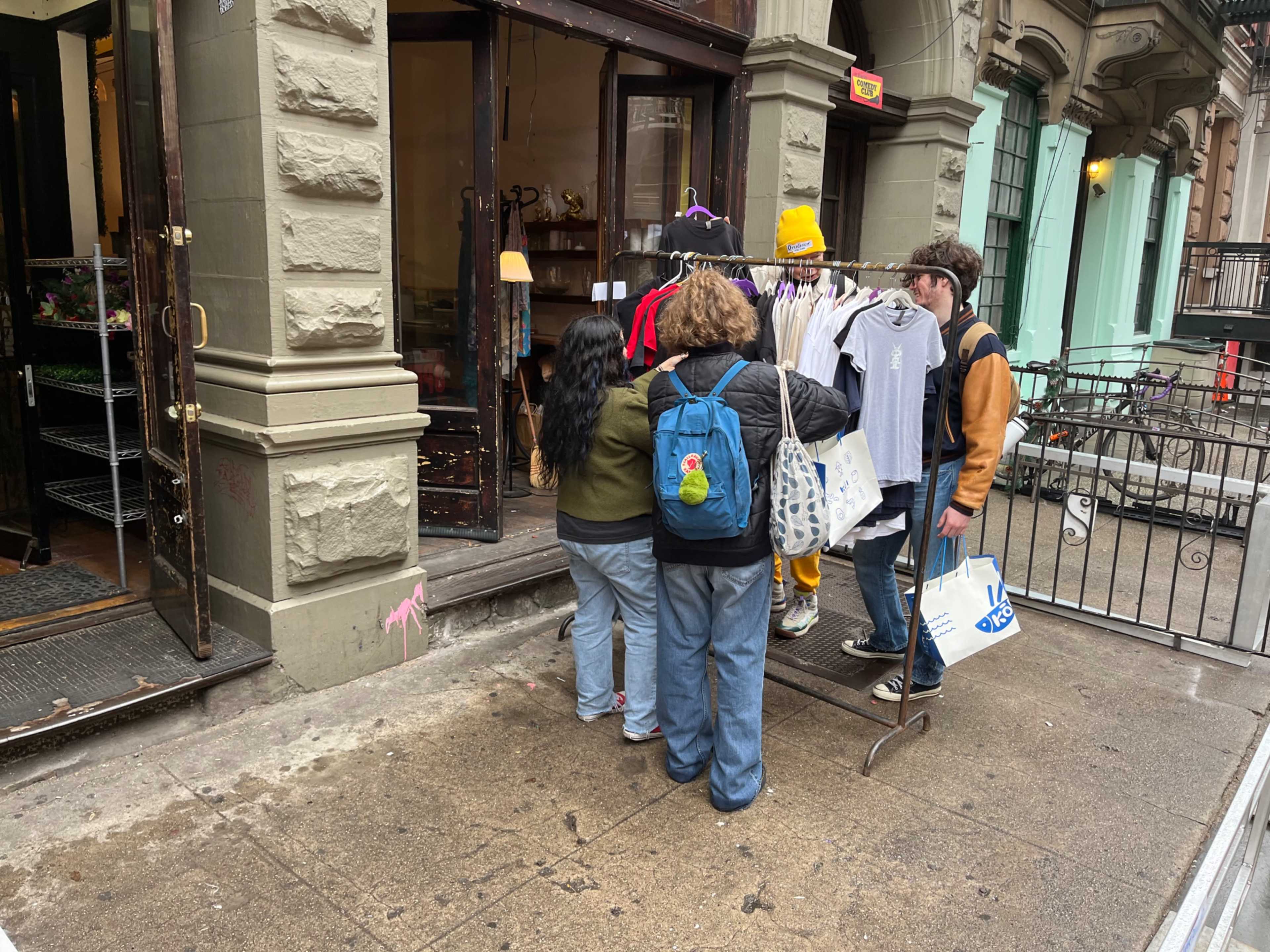 A group of people examines clothing hanging on a rack outside a store in an urban setting.