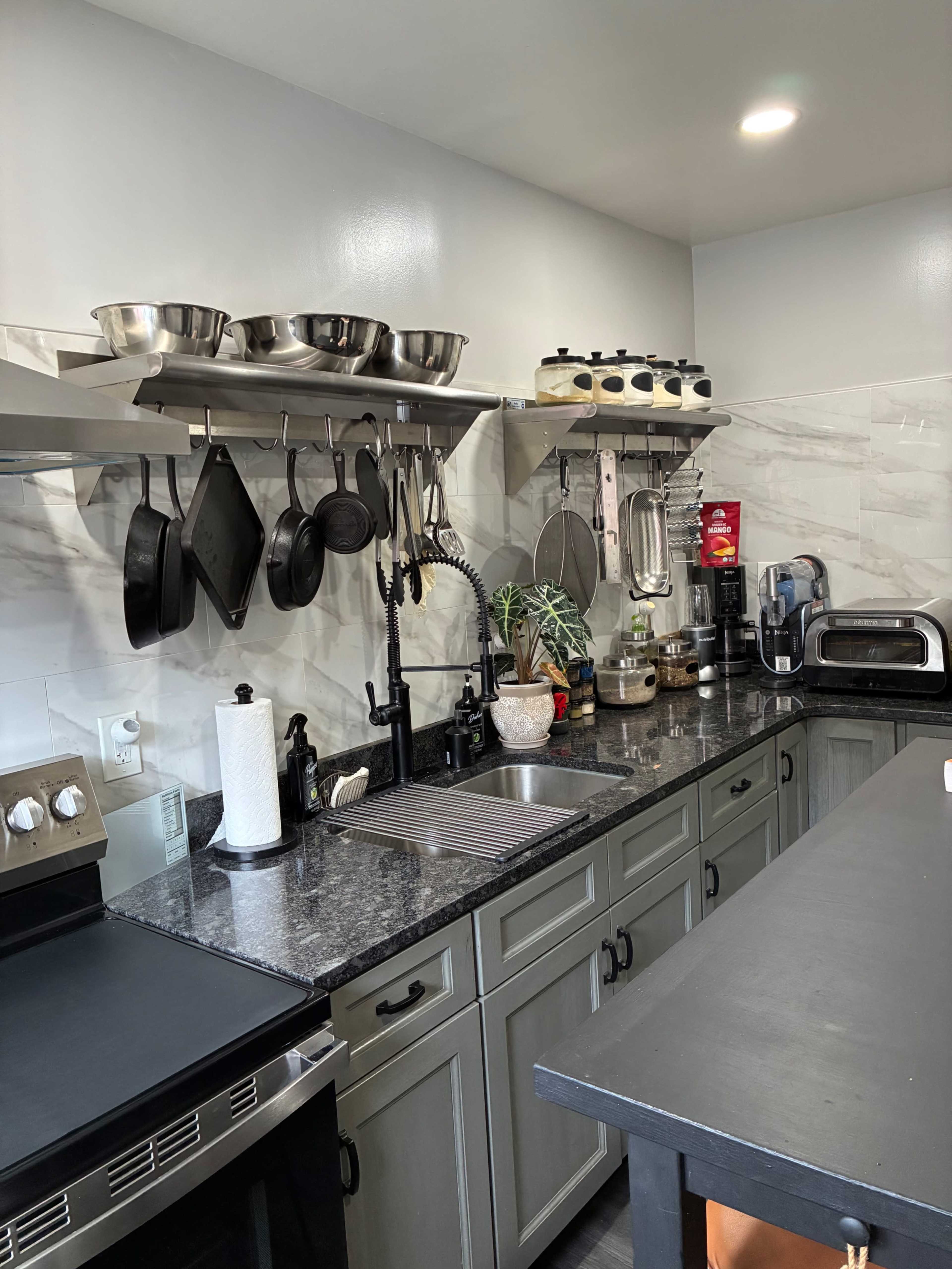 The image shows a modern kitchen with a stainless steel sink, wall-mounted shelves holding cookware, and various appliances on the counter.