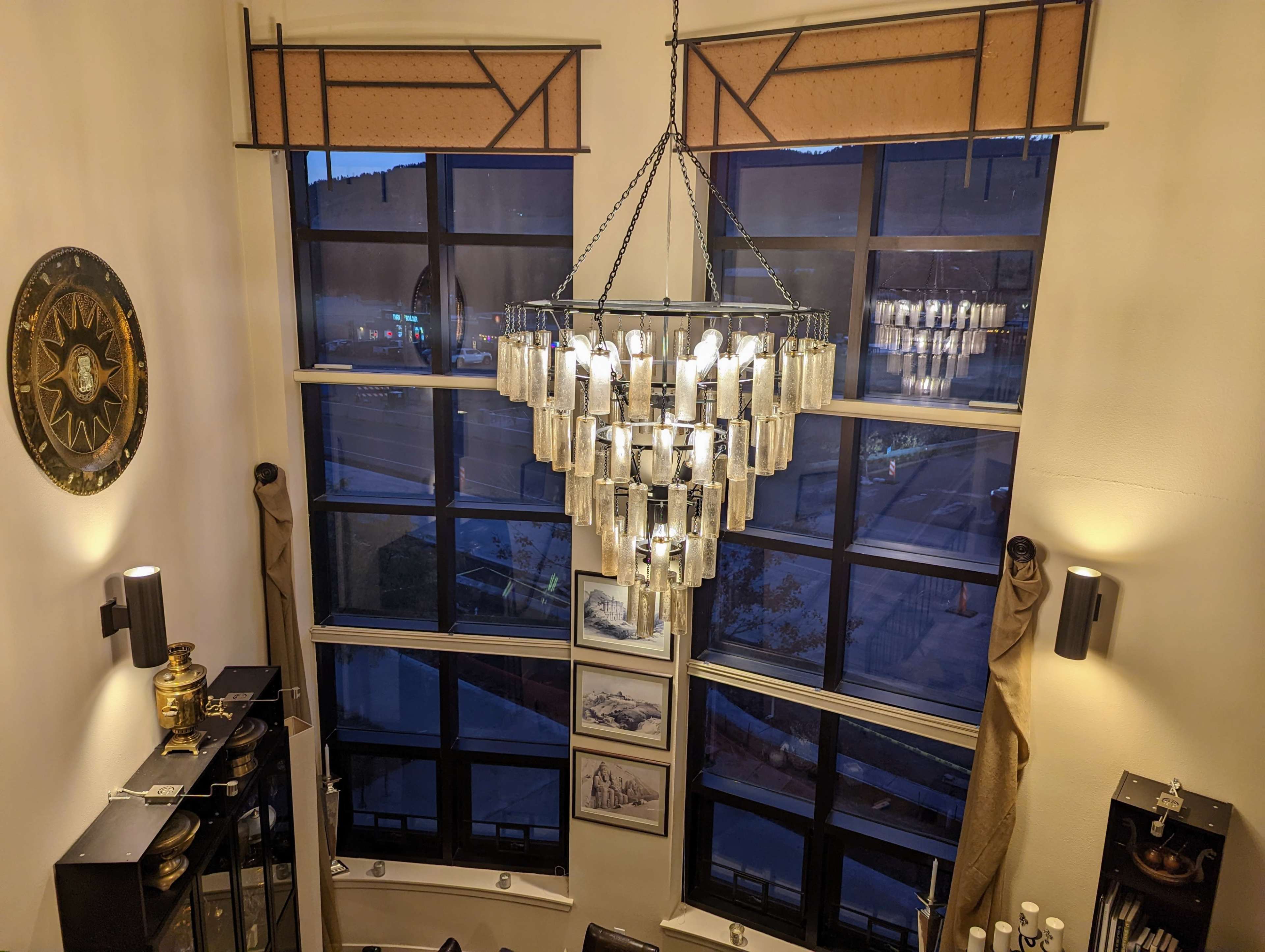 The image shows a well-lit interior with large windows, a modern chandelier hanging from the ceiling, and decorative items on the walls and shelves.