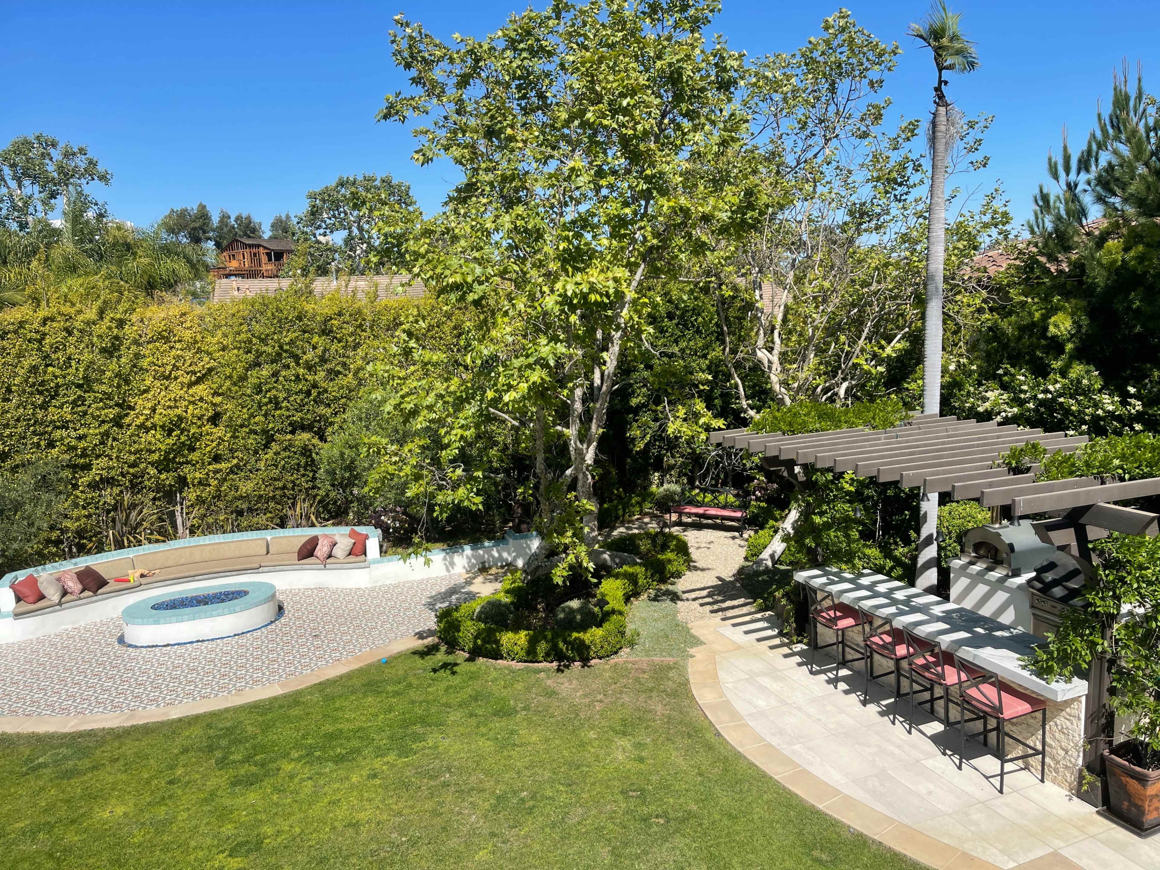 A landscaped outdoor lawn area featuring a patterned stone fire pit surrounded by benches, a pergola with seating, and lush greenery.