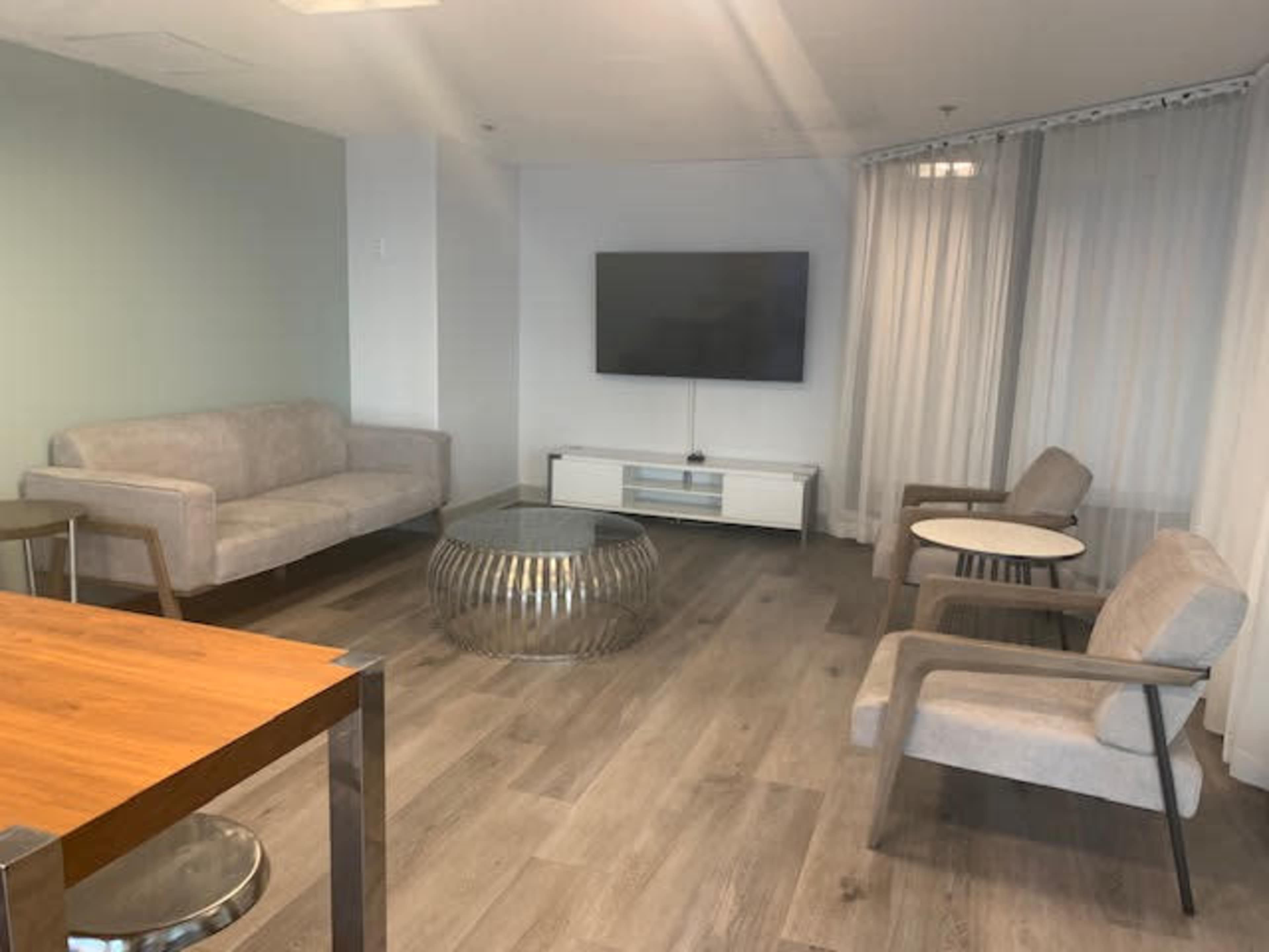 A modern living area with a sofa, two chairs, a glass coffee table, a dining table, and a television mounted on the wall.