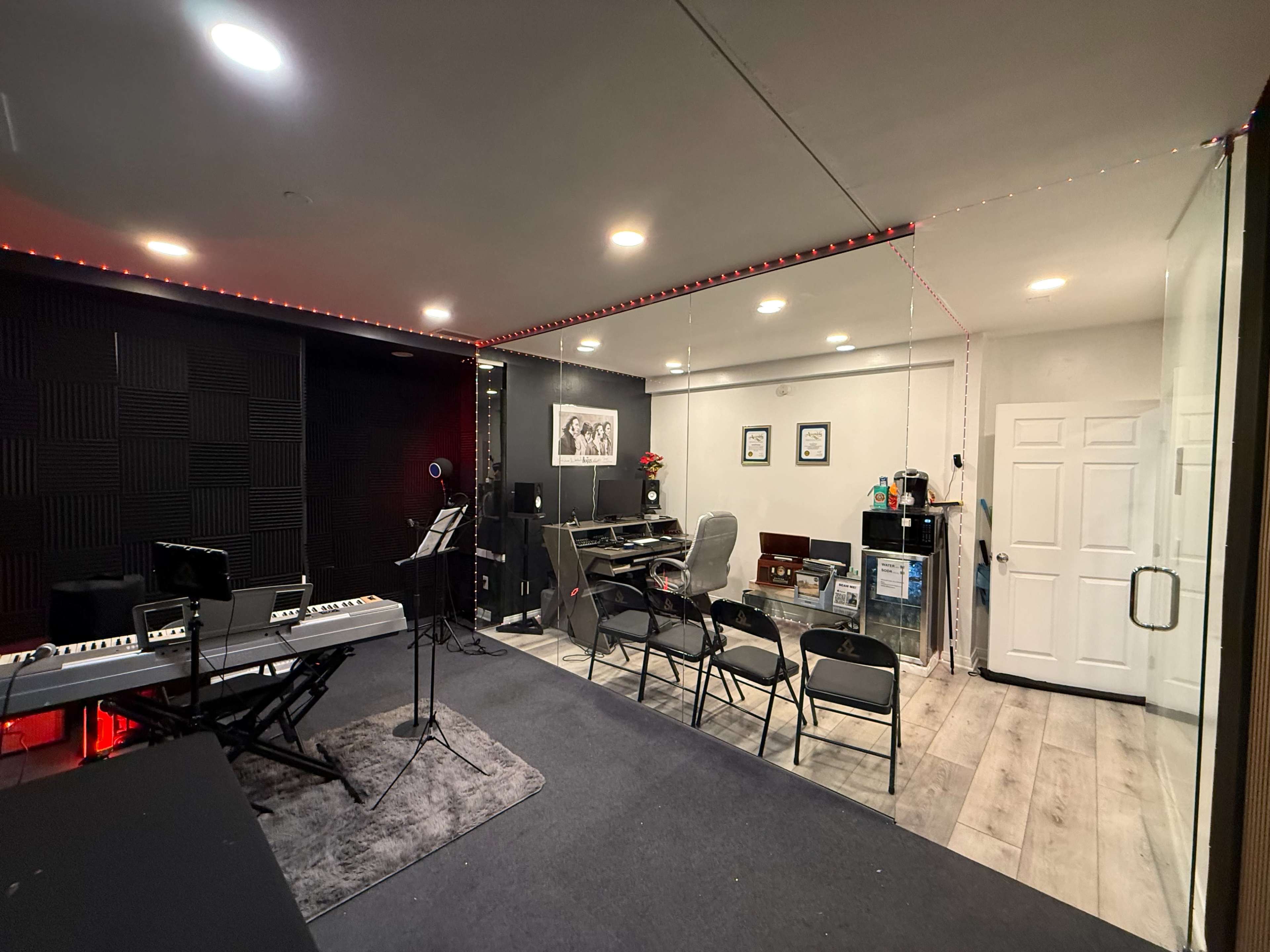 The image shows a music studio featuring soundproofed walls, a row of chairs, and equipment including a keyboard, computer workstation, and glass door.