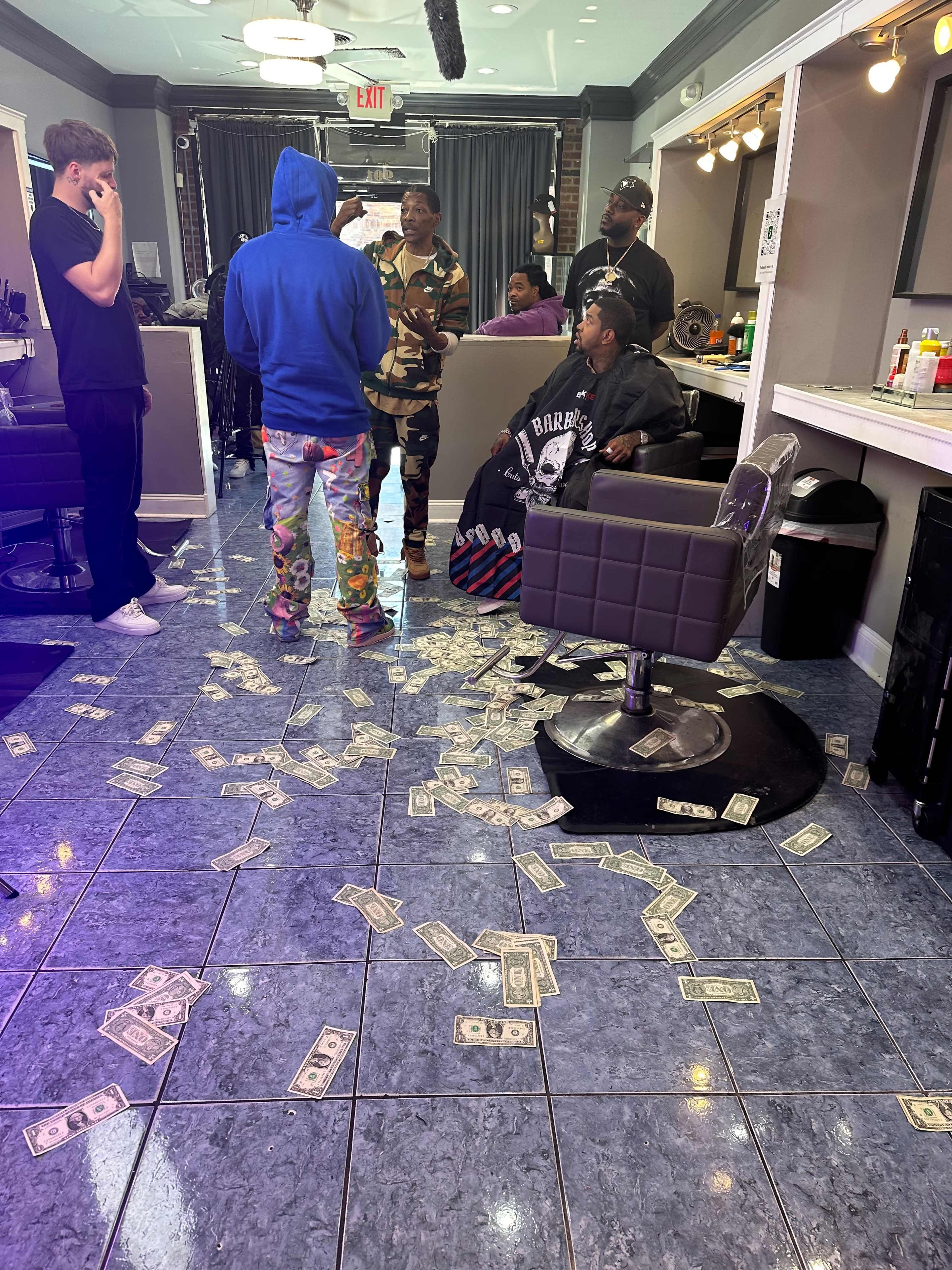 A barbershop interior features several people, with money scattered across the floor and barbershop chairs.