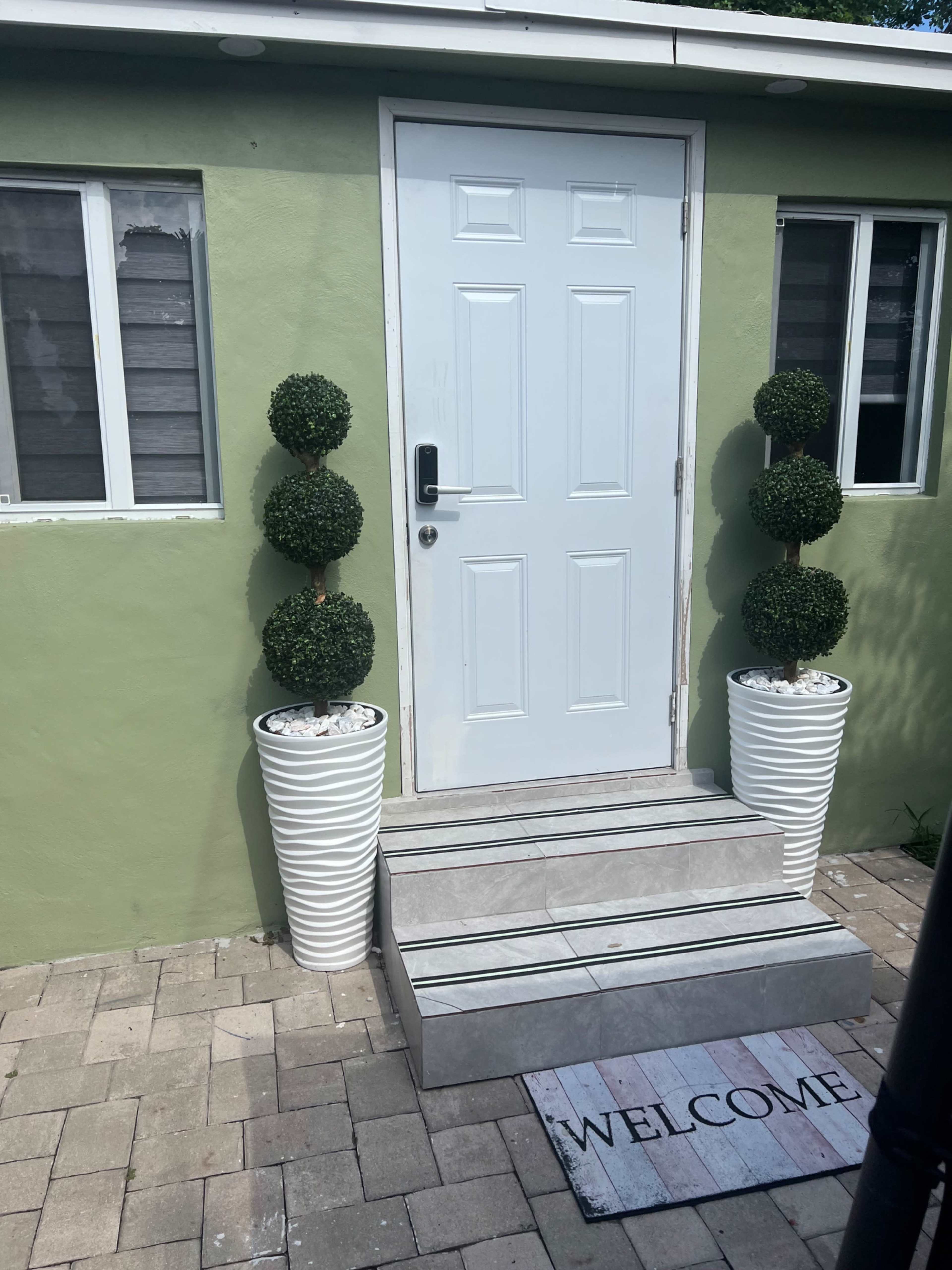 A green building with a white door and potted topiary plants on either side of a set of steps leading up to the entrance.