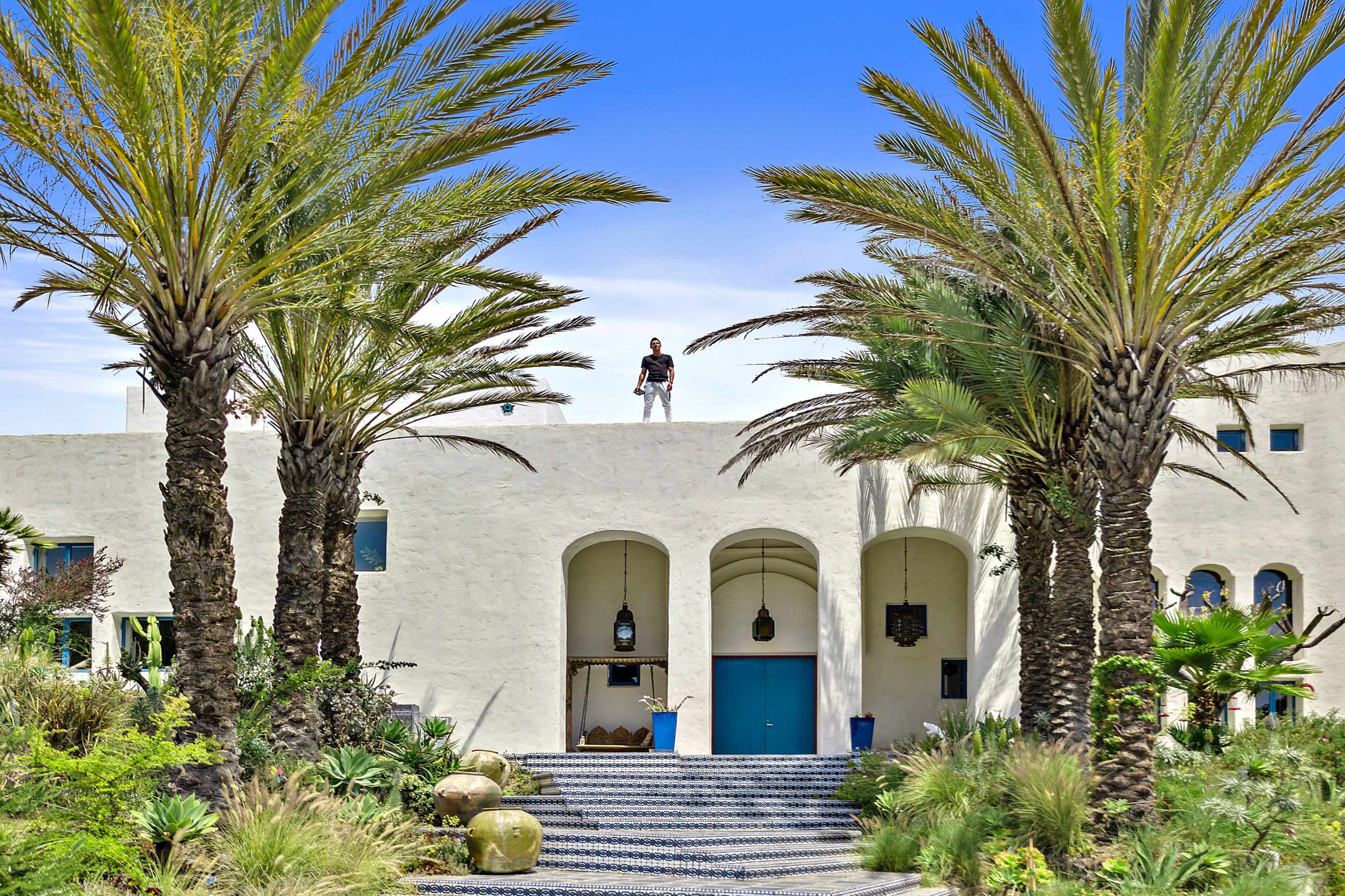 A person stands on the roof of a white building surrounded by palm trees and landscaped greenery.
