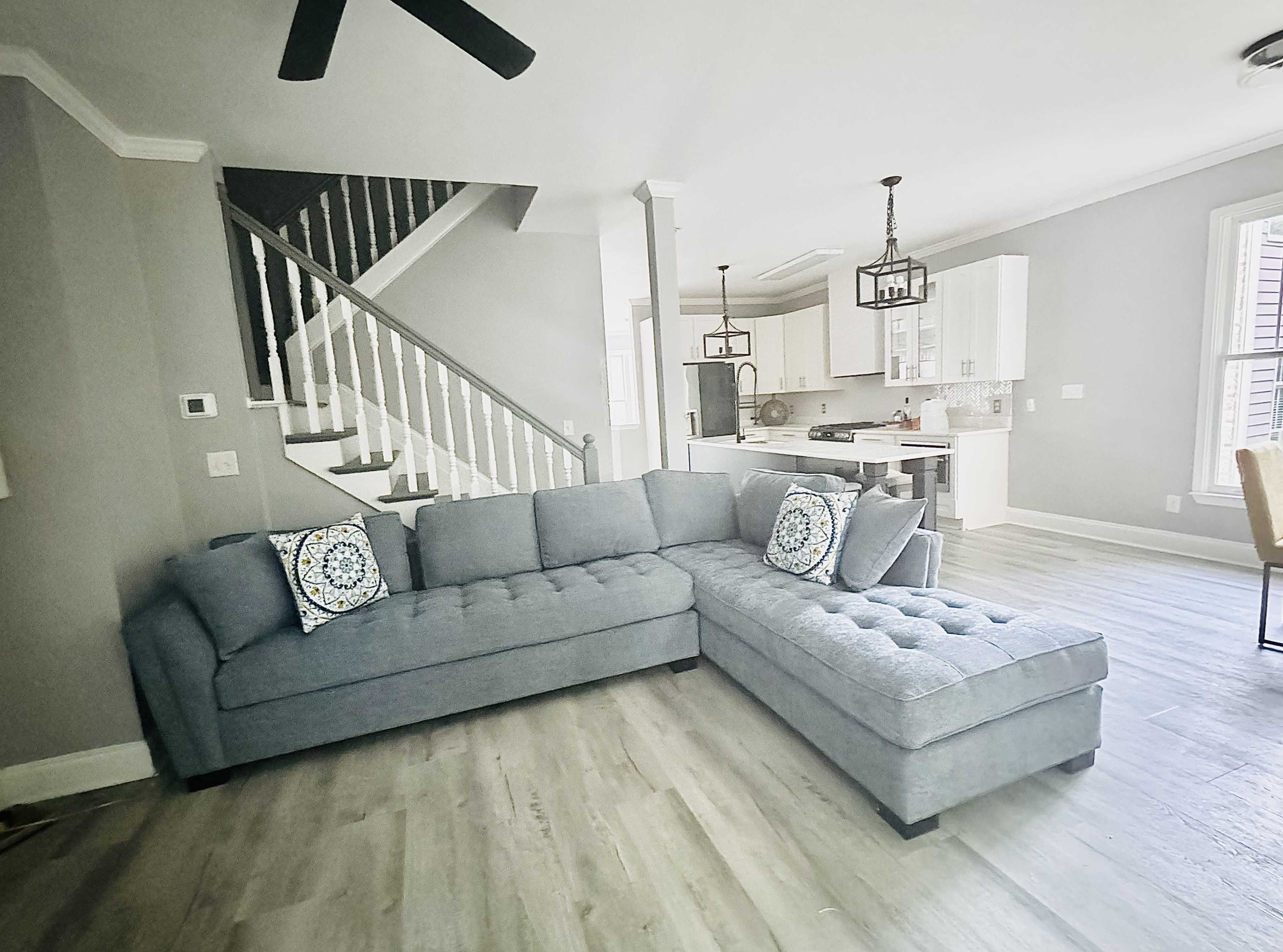 The image shows a gray sectional sofa with decorative pillows positioned in a modern living room that leads to a kitchen with white cabinetry and a staircase in the background.