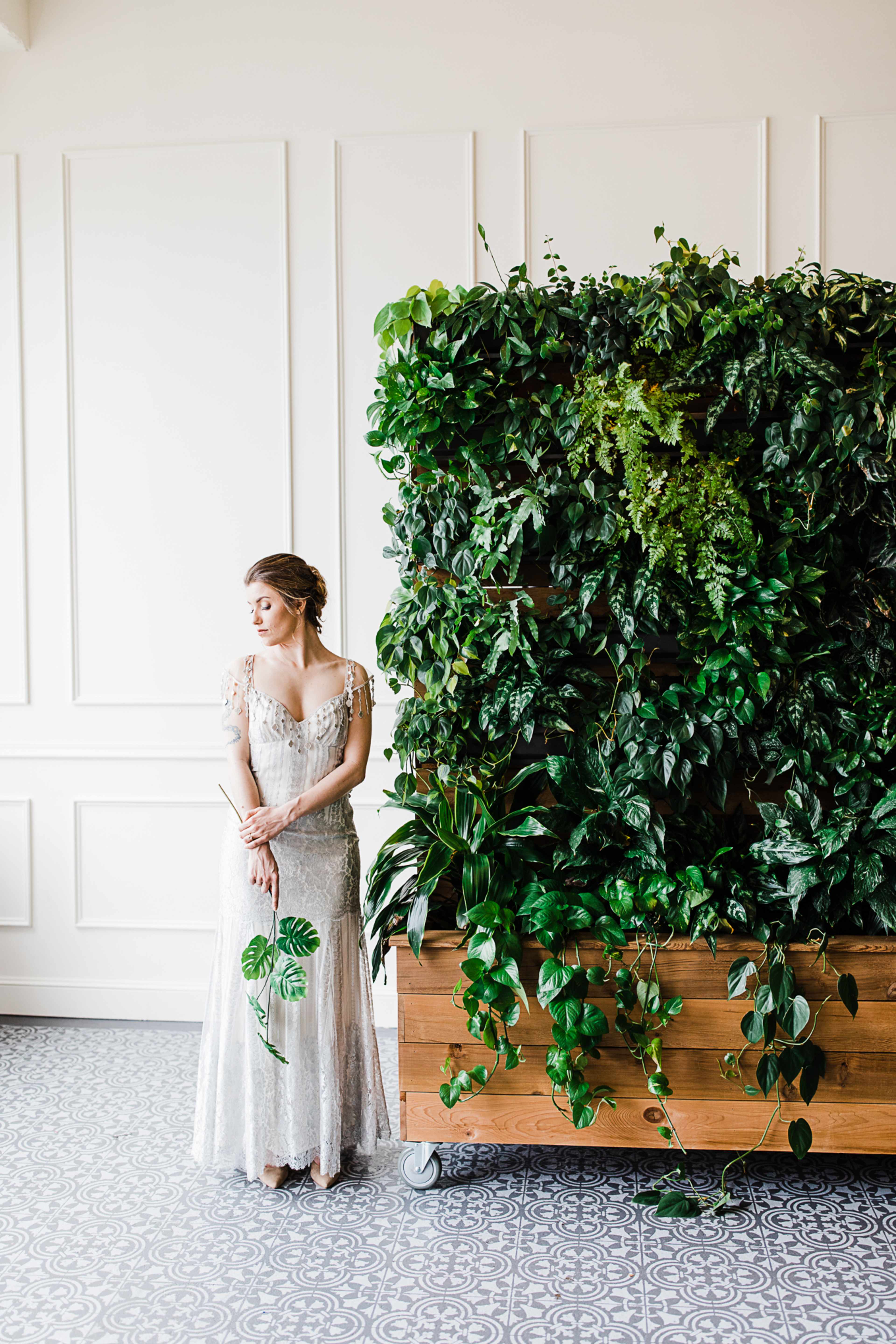 A woman in a silver gown stands beside a tall, lush greenery arrangement against a white wall.
