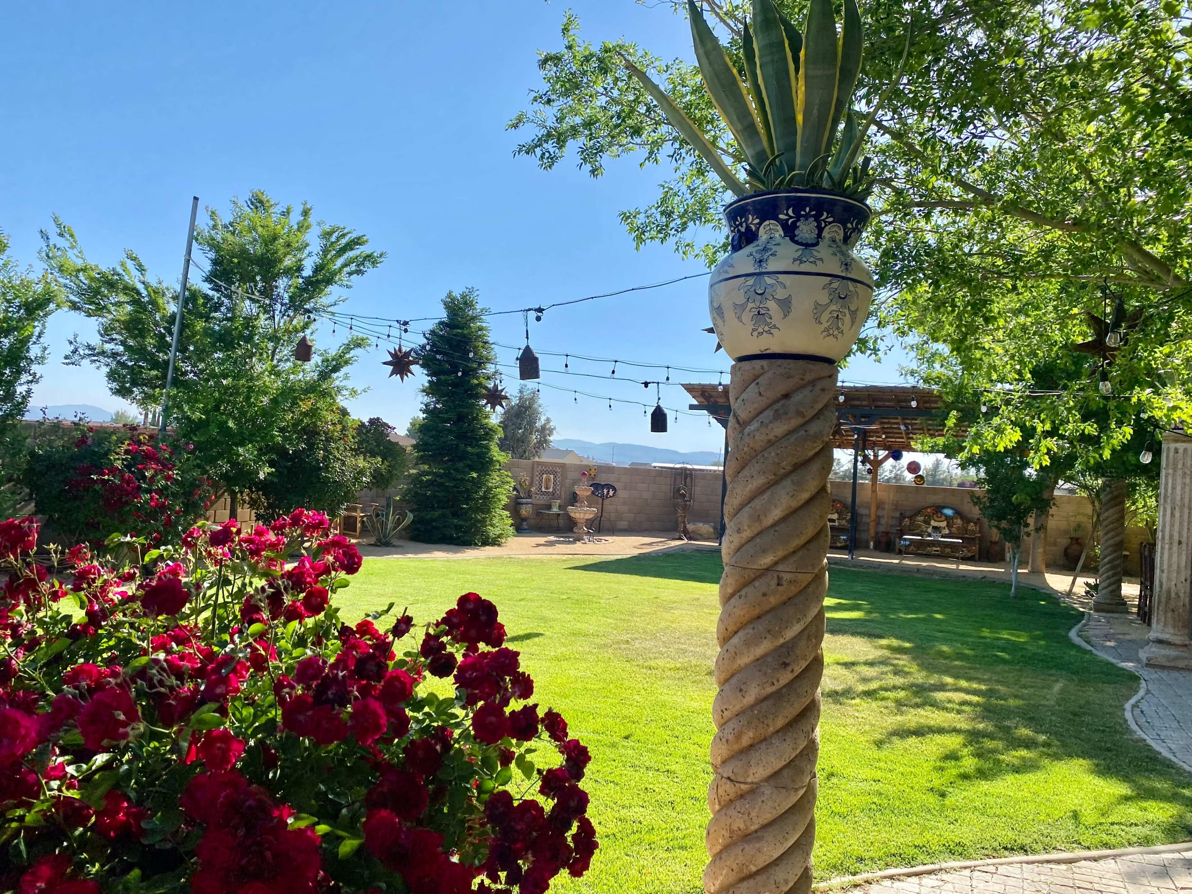 A landscaped garden featuring a flower bed of red roses, a tall column adorned with a potted plant, and a grassy area surrounded by trees and decorative elements.