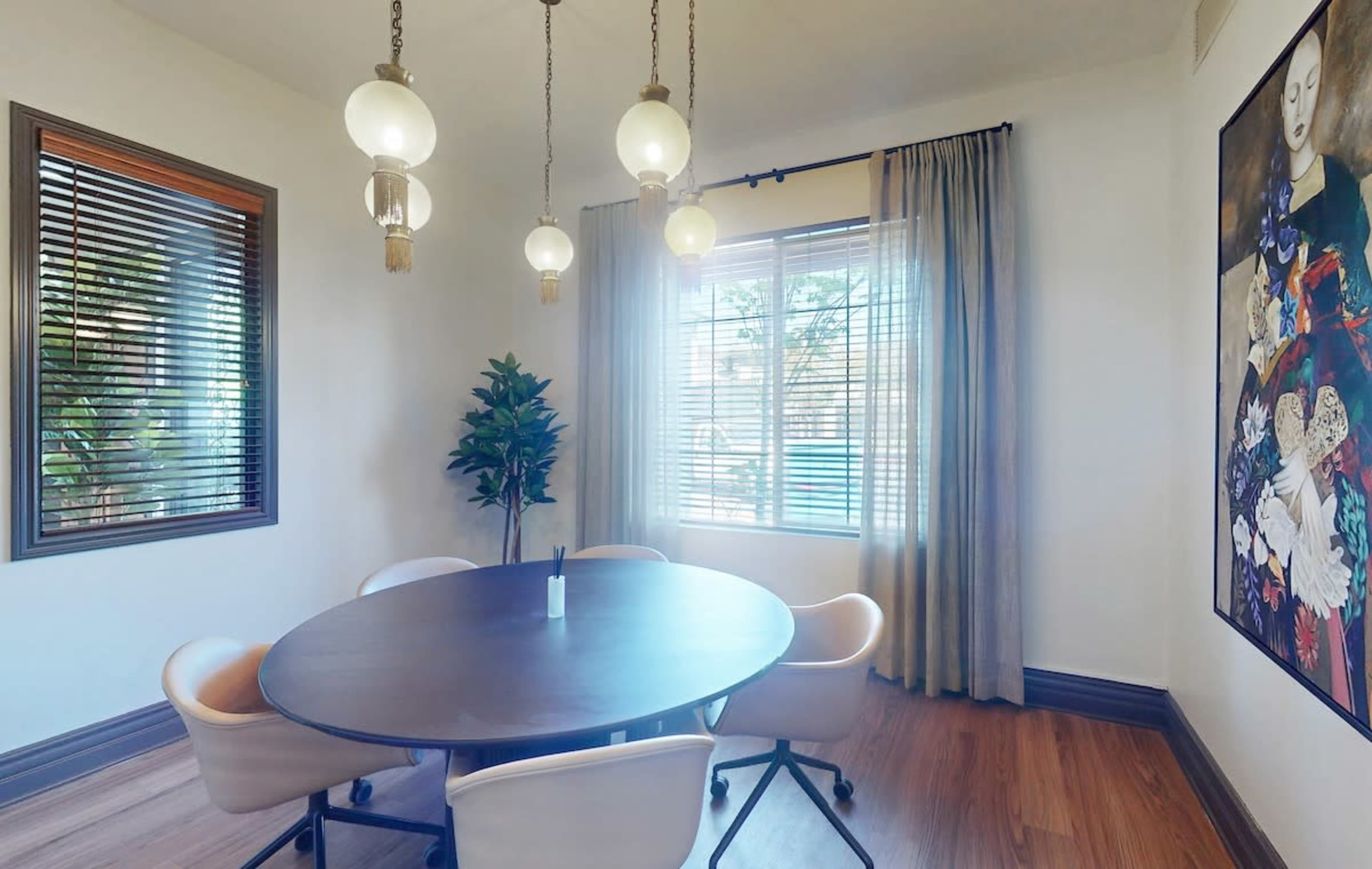 A modern dining area features a round table surrounded by four white chairs, illuminated by pendant lights, with a large window and a painting on the wall.