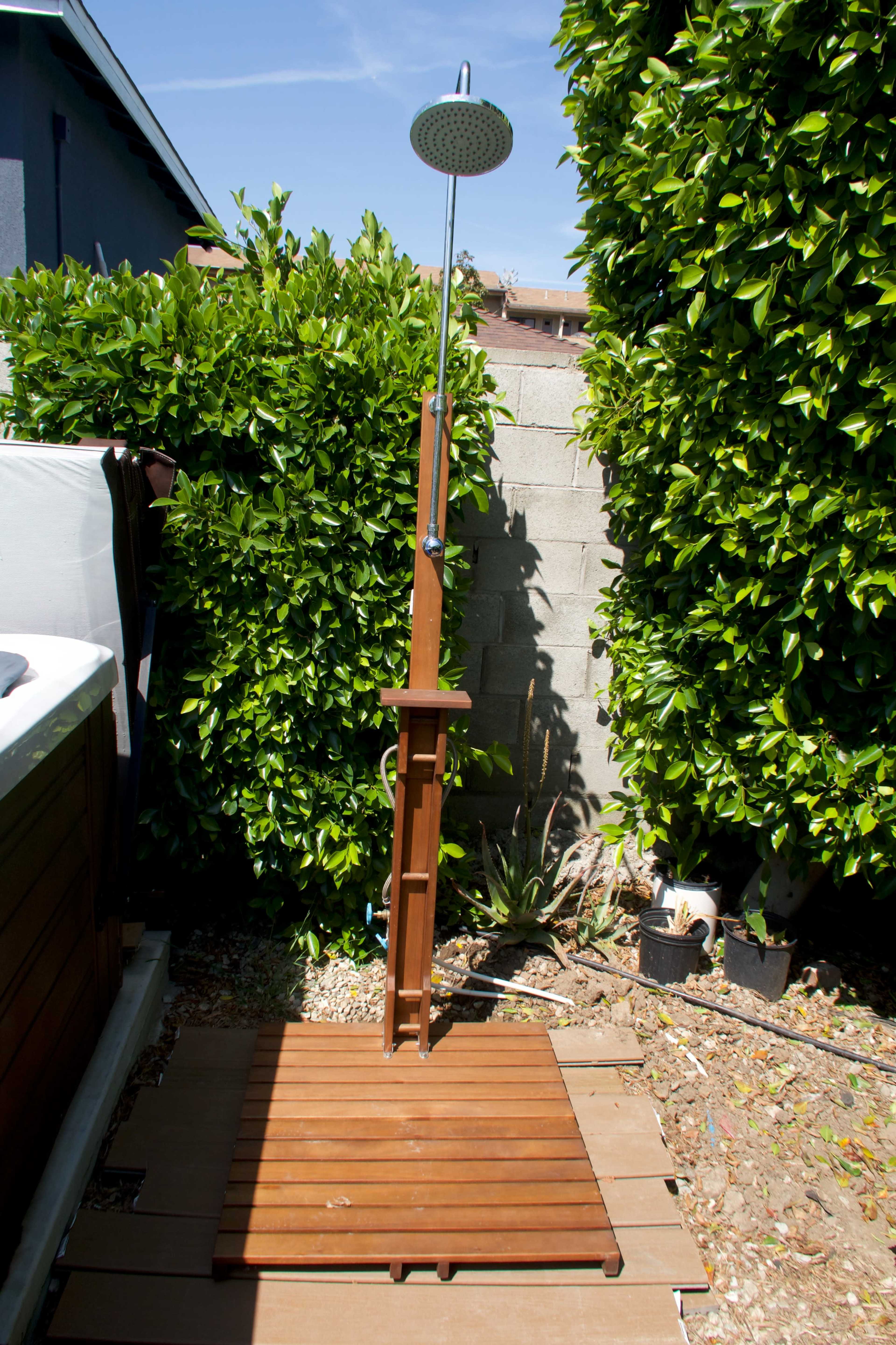 An outdoor shower made of wood, positioned on a wooden deck beside a privacy fence and surrounded by leafy green plants.