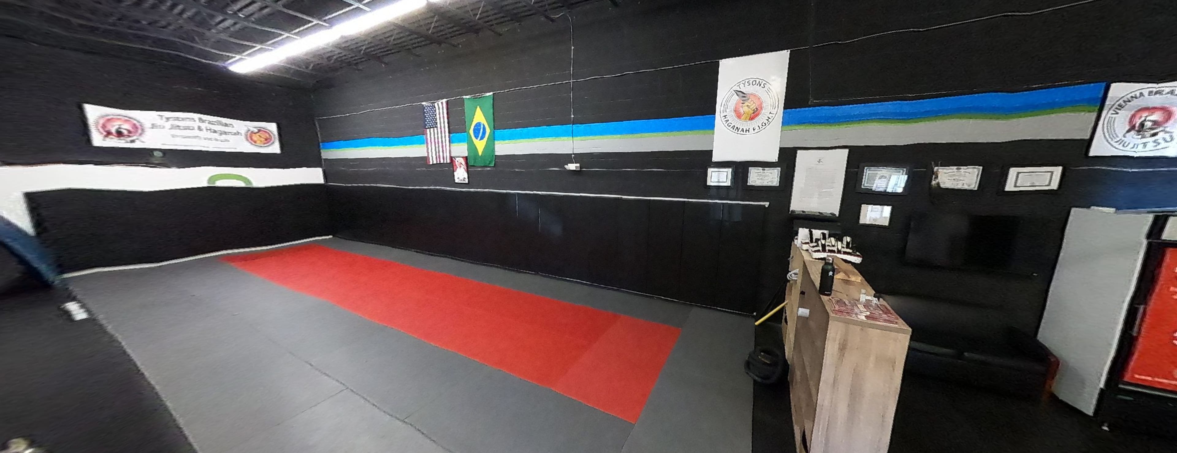 A martial arts training space with red and gray mats, black walls adorned with flags, and a seating area.