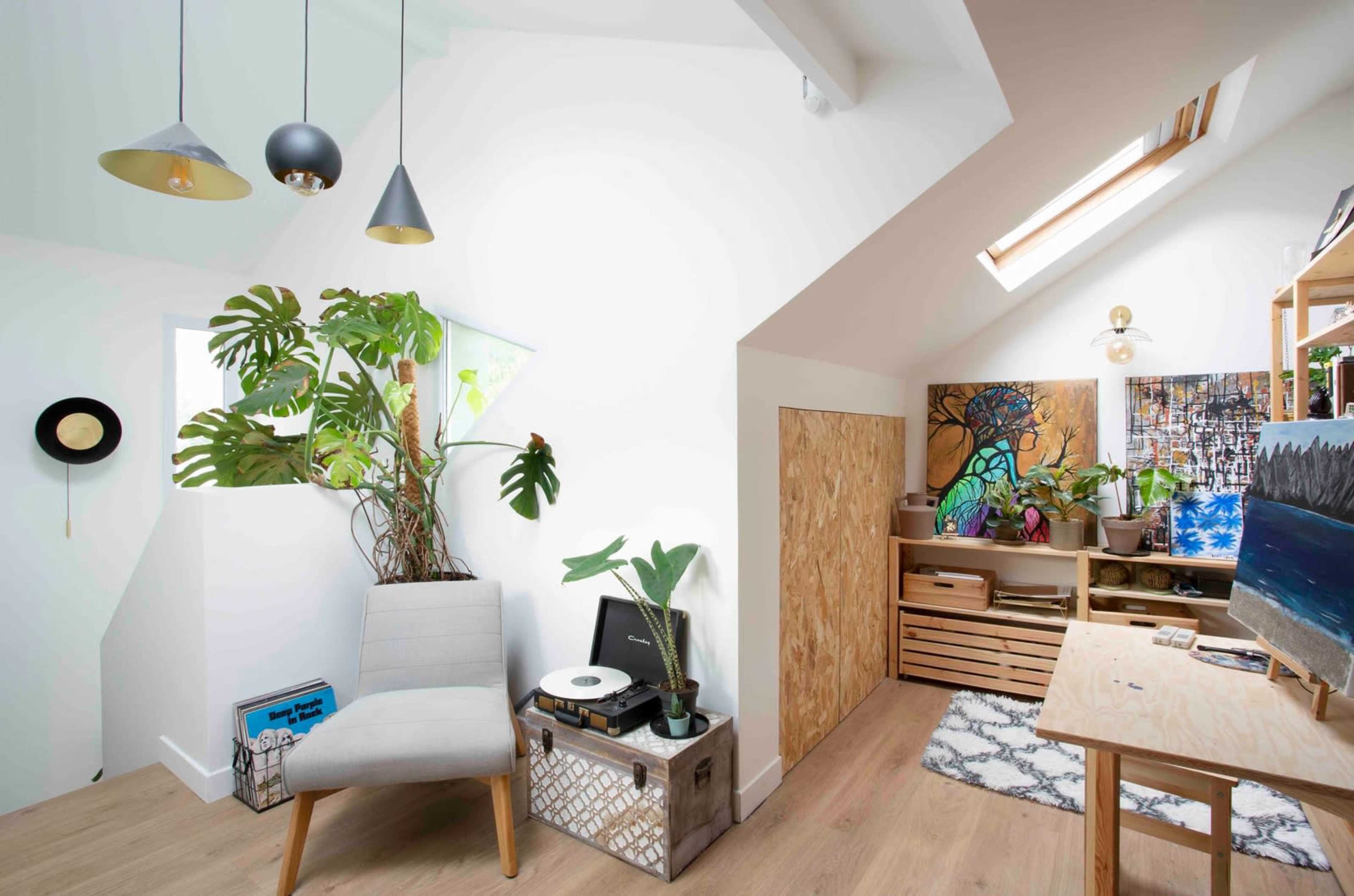 The image shows a well-lit room with a chair, a record player, shelves filled with plants and art supplies, and a skylight above.