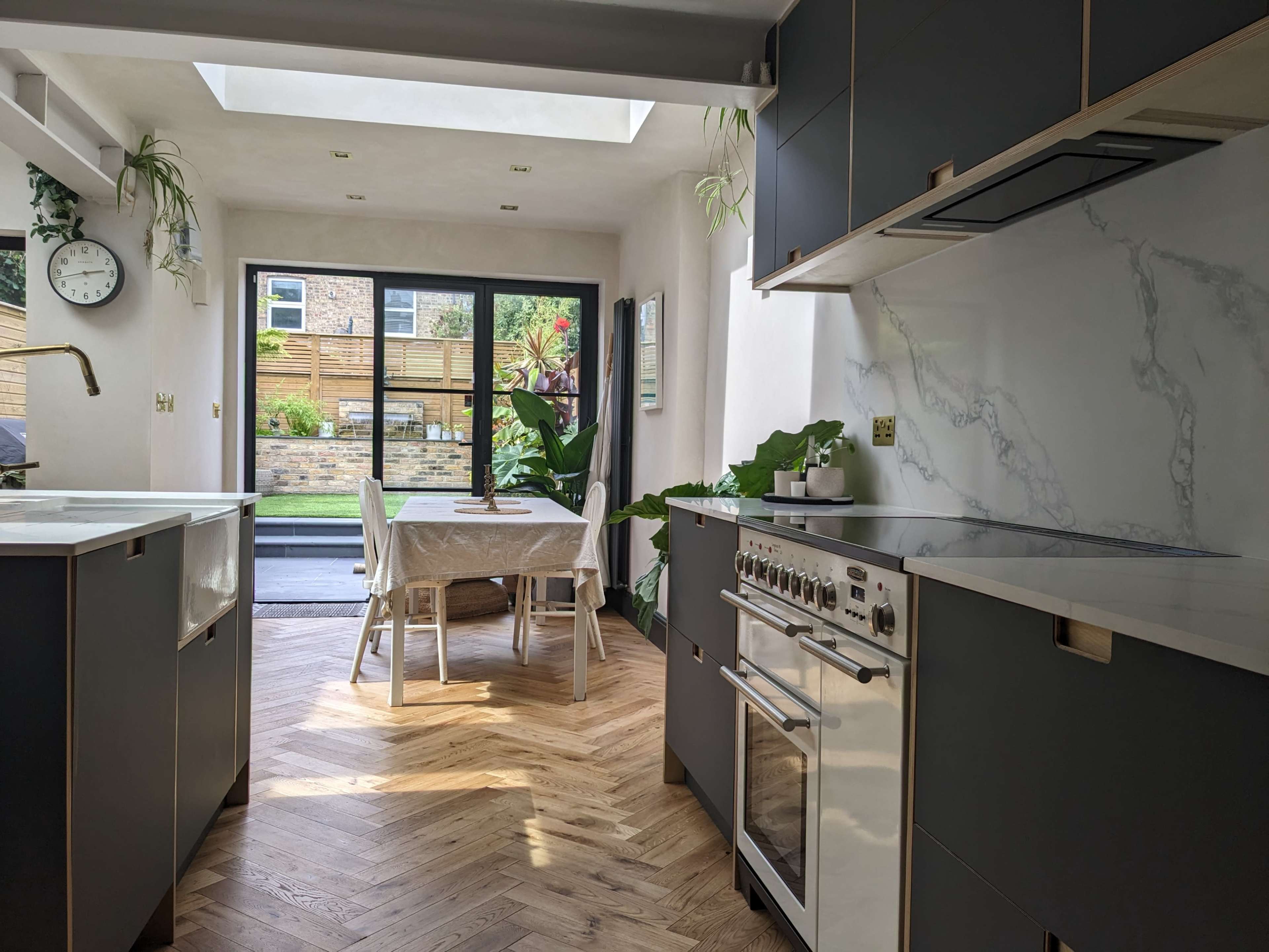 A modern kitchen with dark cabinetry, a marble countertop, and a view of a dining area and garden through sliding glass doors.