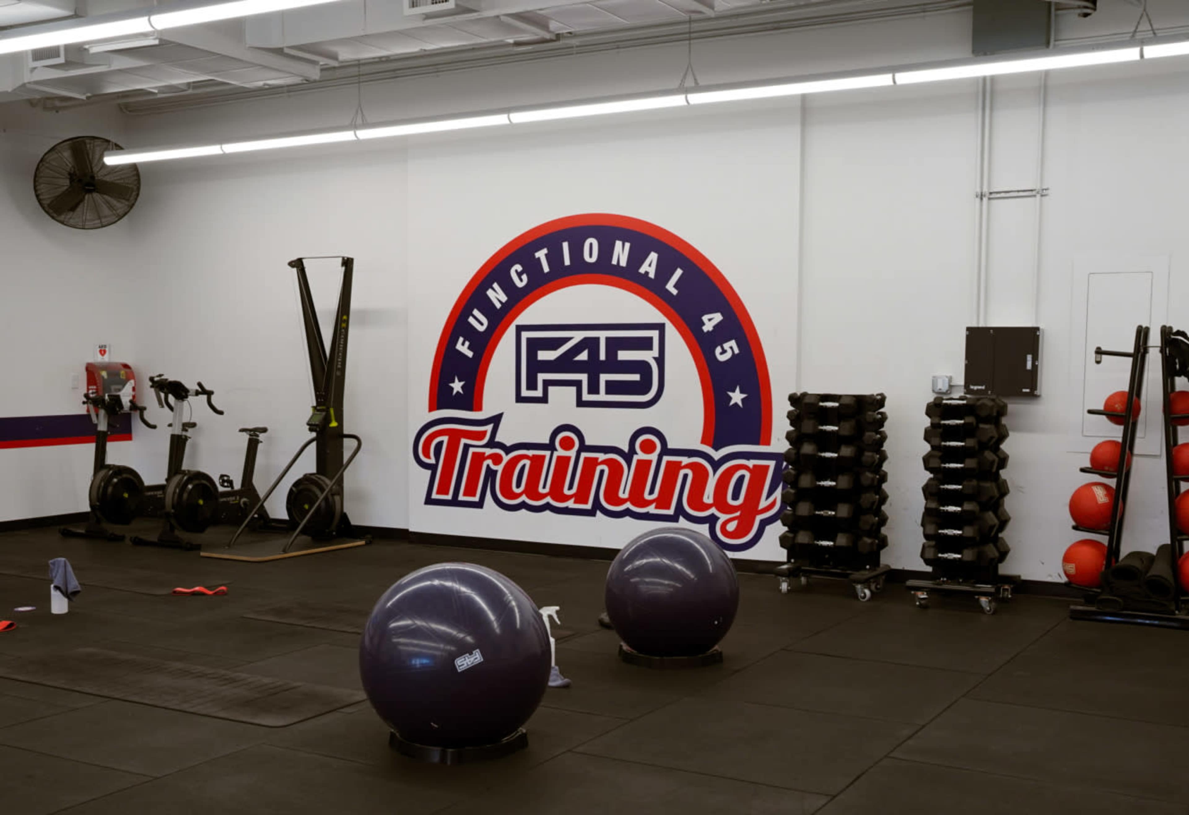 The image shows a fitness training facility featuring equipment such as exercise balls, dumbbells, and a large wall with the "F45 Training" logo.