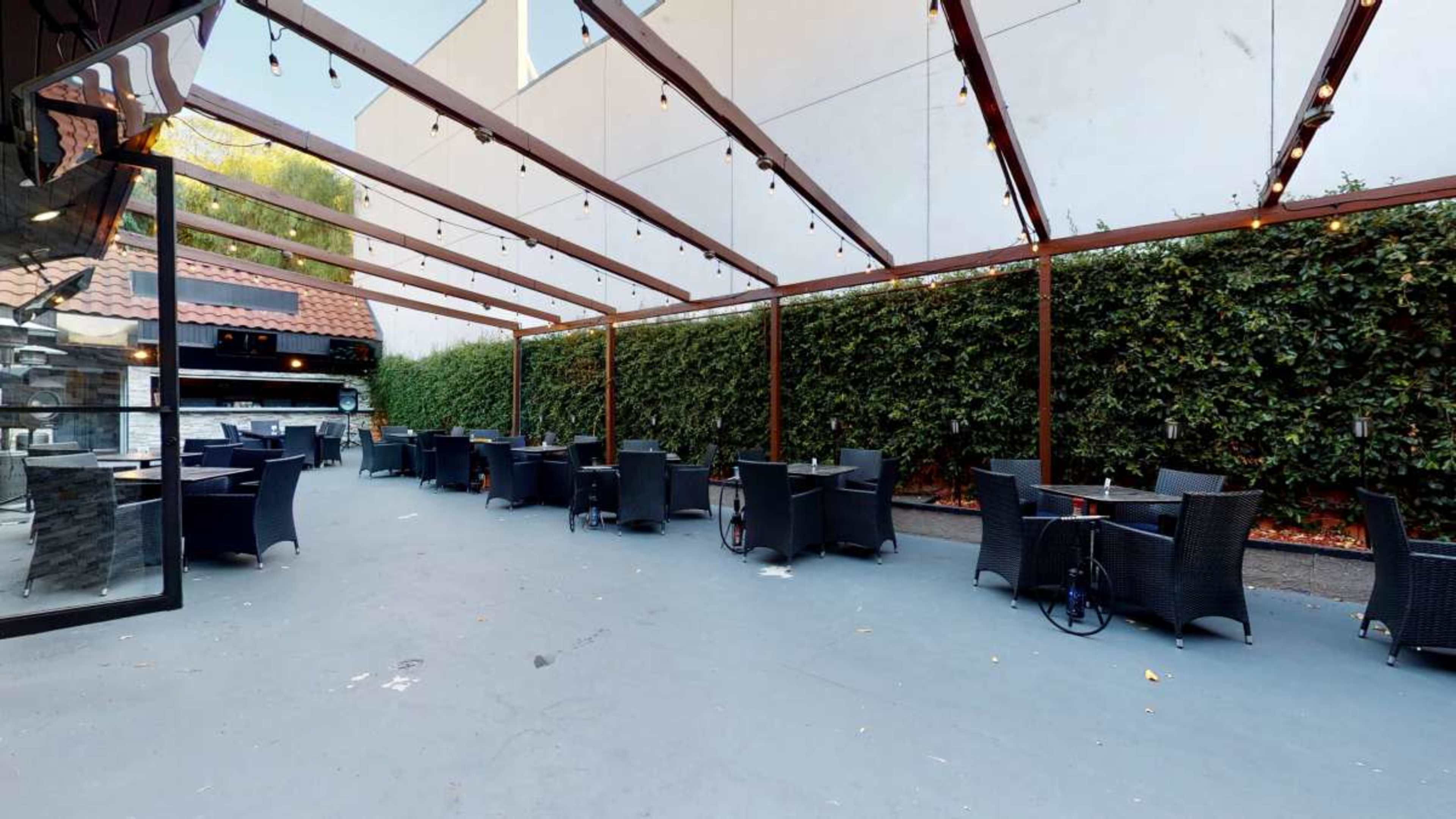 The image shows an outdoor dining area with black wicker chairs and tables arranged under a wooden canopy strung with lights.