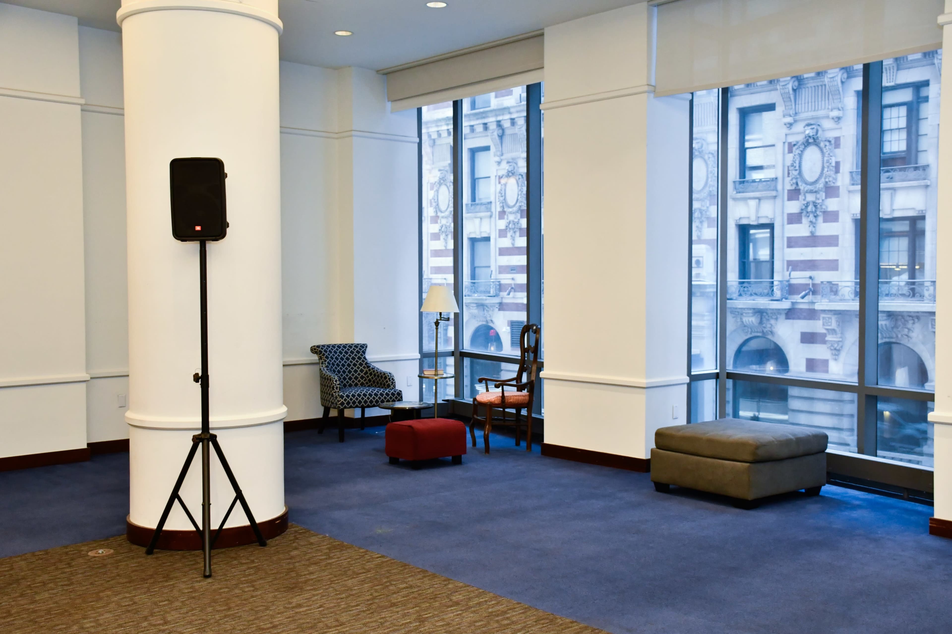 A modern lobby area features a speaker on a stand, two chairs, a lamp, and a small ottoman near large windows that showcase an urban view.