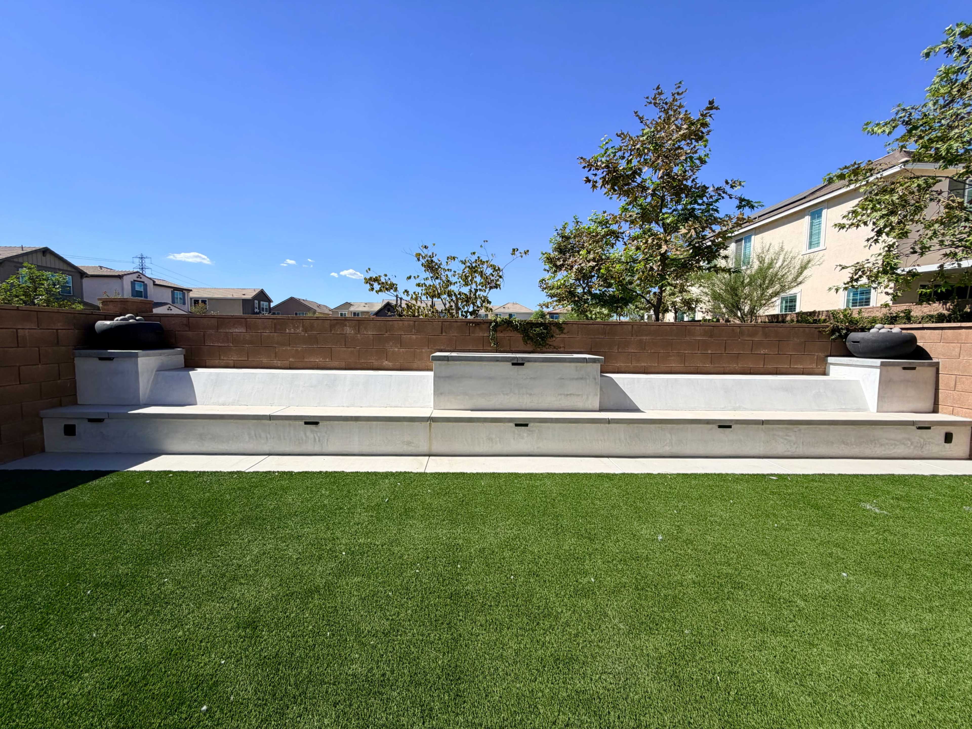 The image shows a backyard seating area made of concrete with a planter in the center, surrounded by artificial grass and residential buildings in the background.