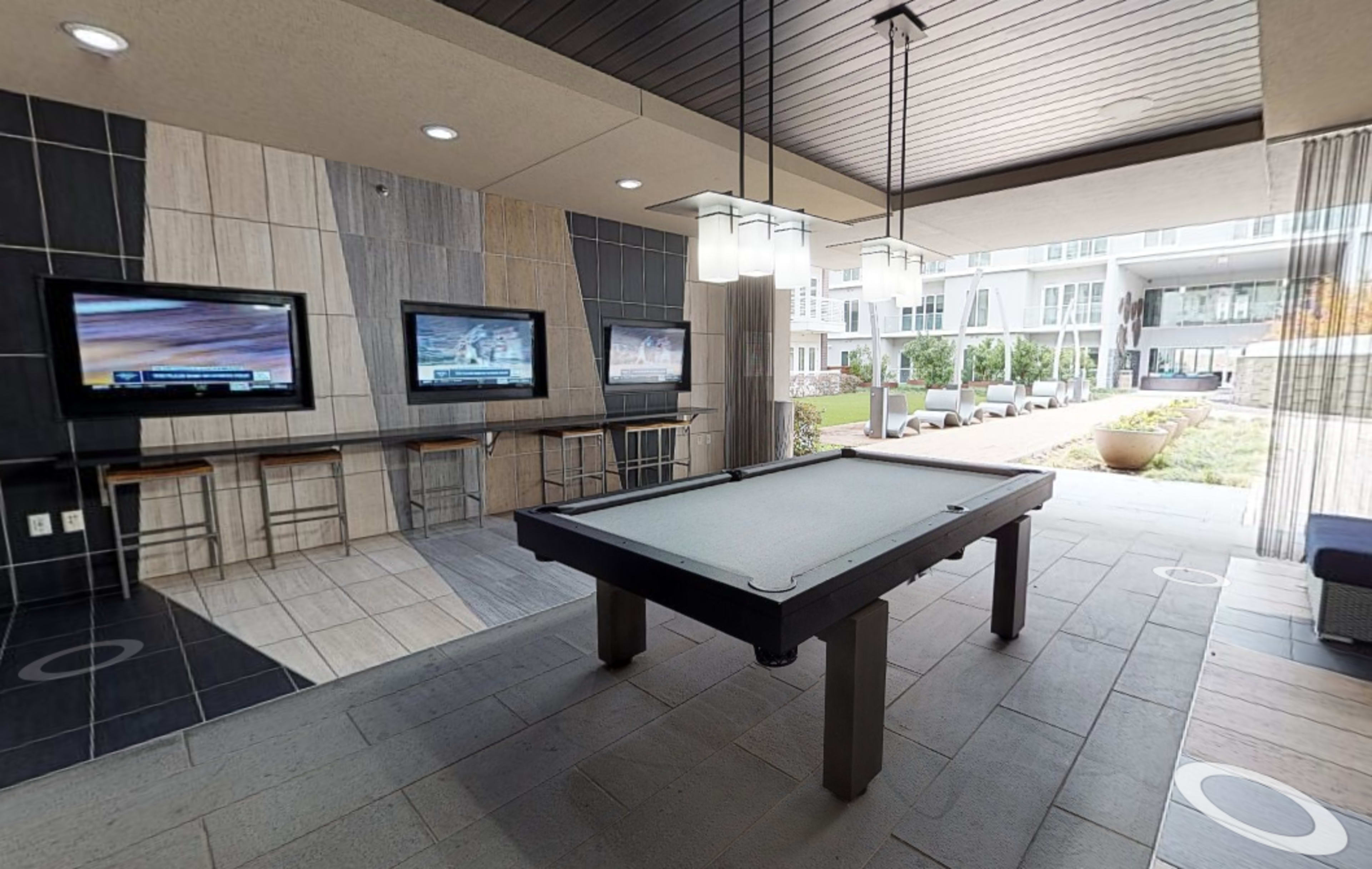 A modern lounge area featuring a pool table, two wall-mounted televisions, and a view of an outdoor seating area.