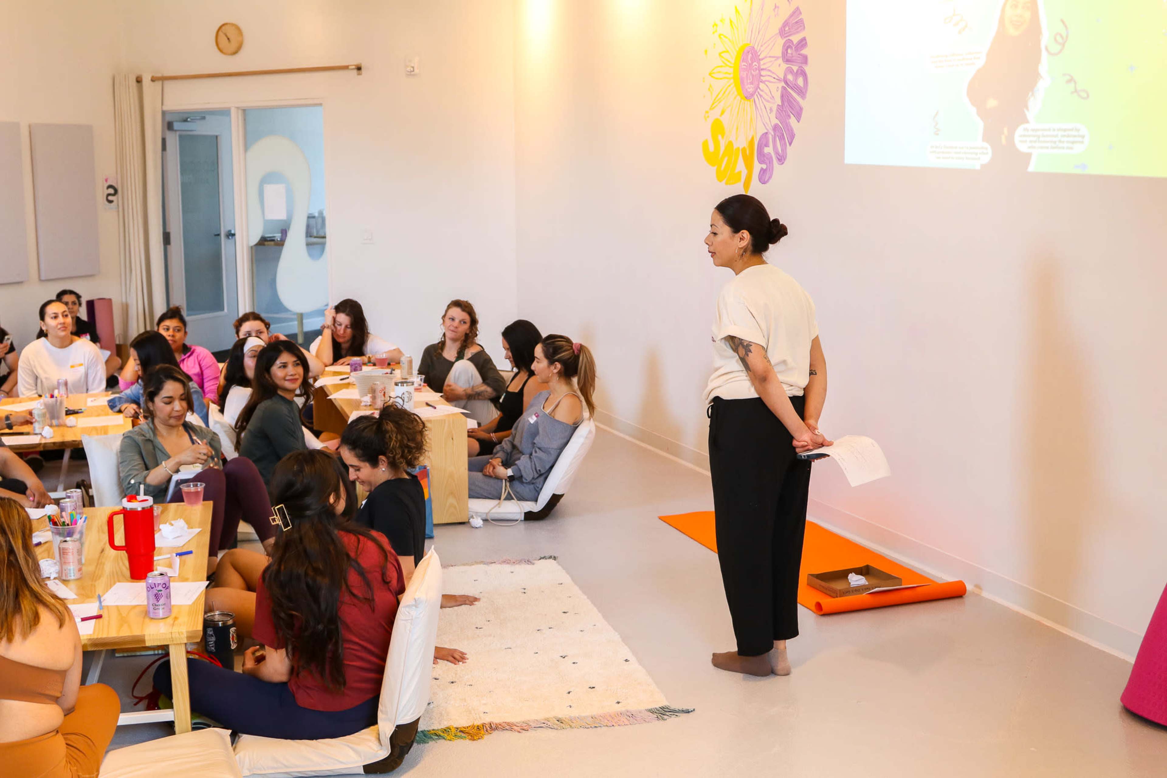 A group of people sits at tables in a brightly lit room, while a woman stands at the front, speaking to them as a presentation is displayed on the wall.