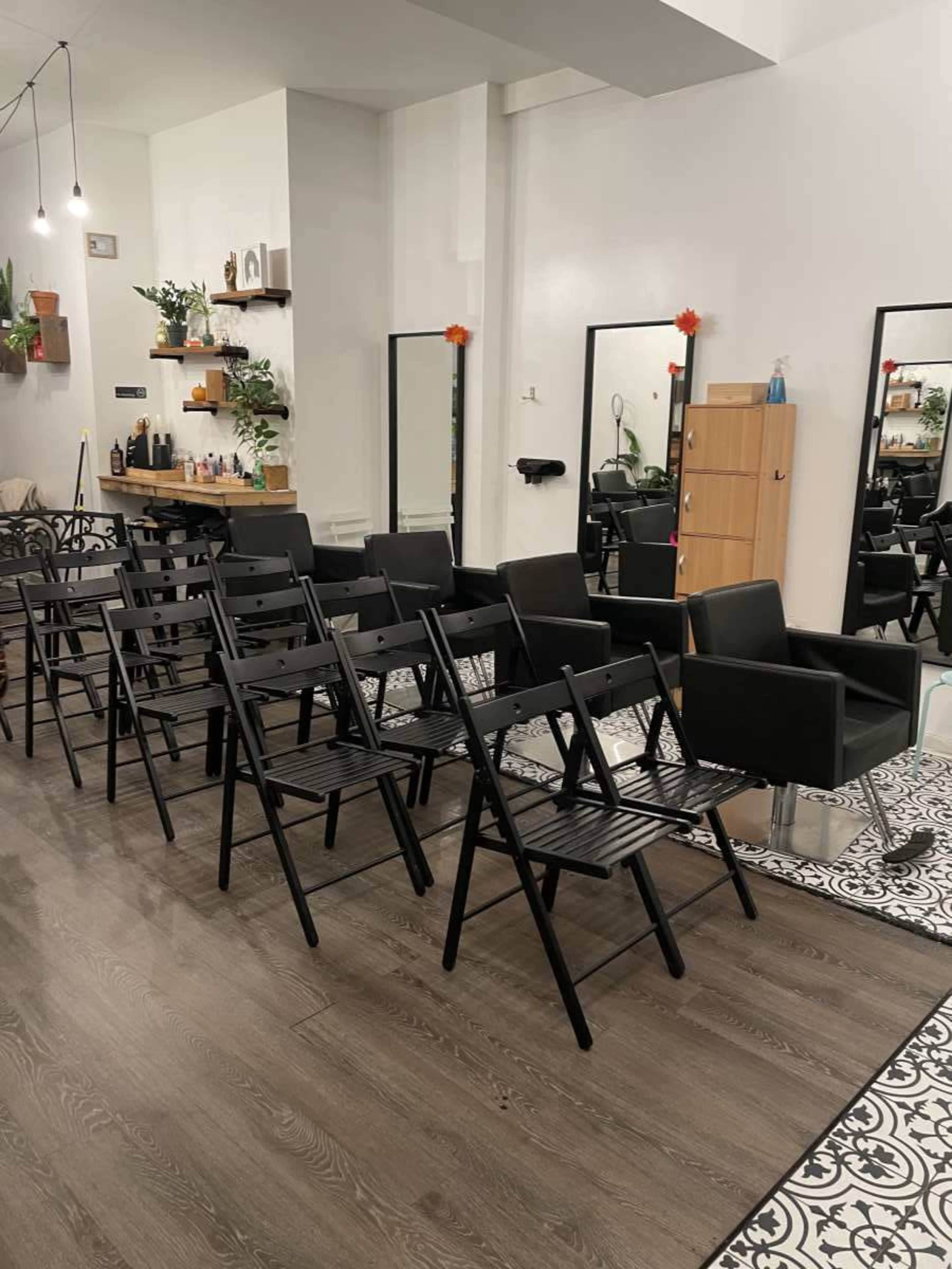 The image shows a well-lit room with a row of black folding chairs facing a mirror and a hairstyling chair, situated on a patterned floor.