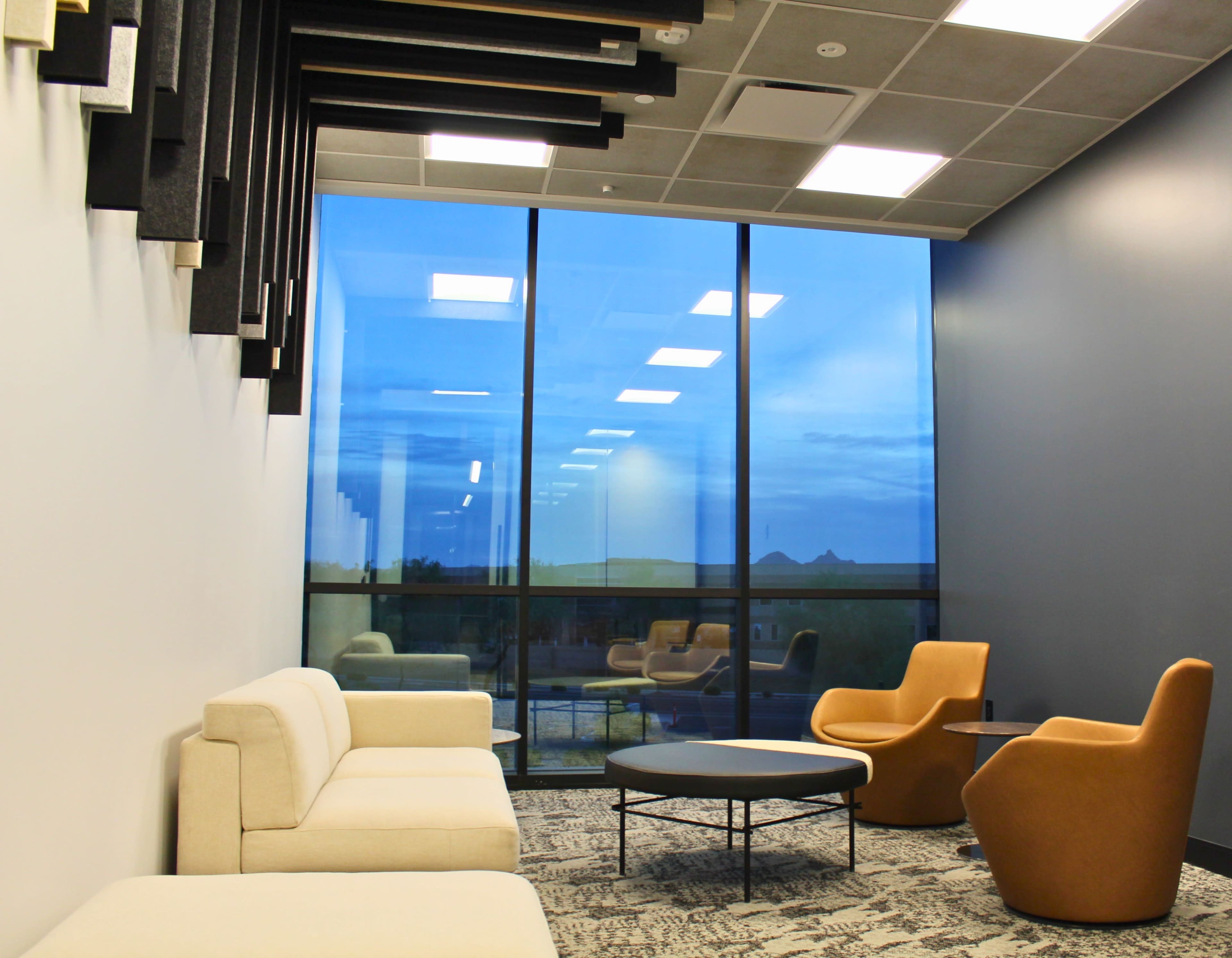 The image shows a modern lounge area with a white sofa, orange chairs, a round table, and large windows reflecting a twilight sky.