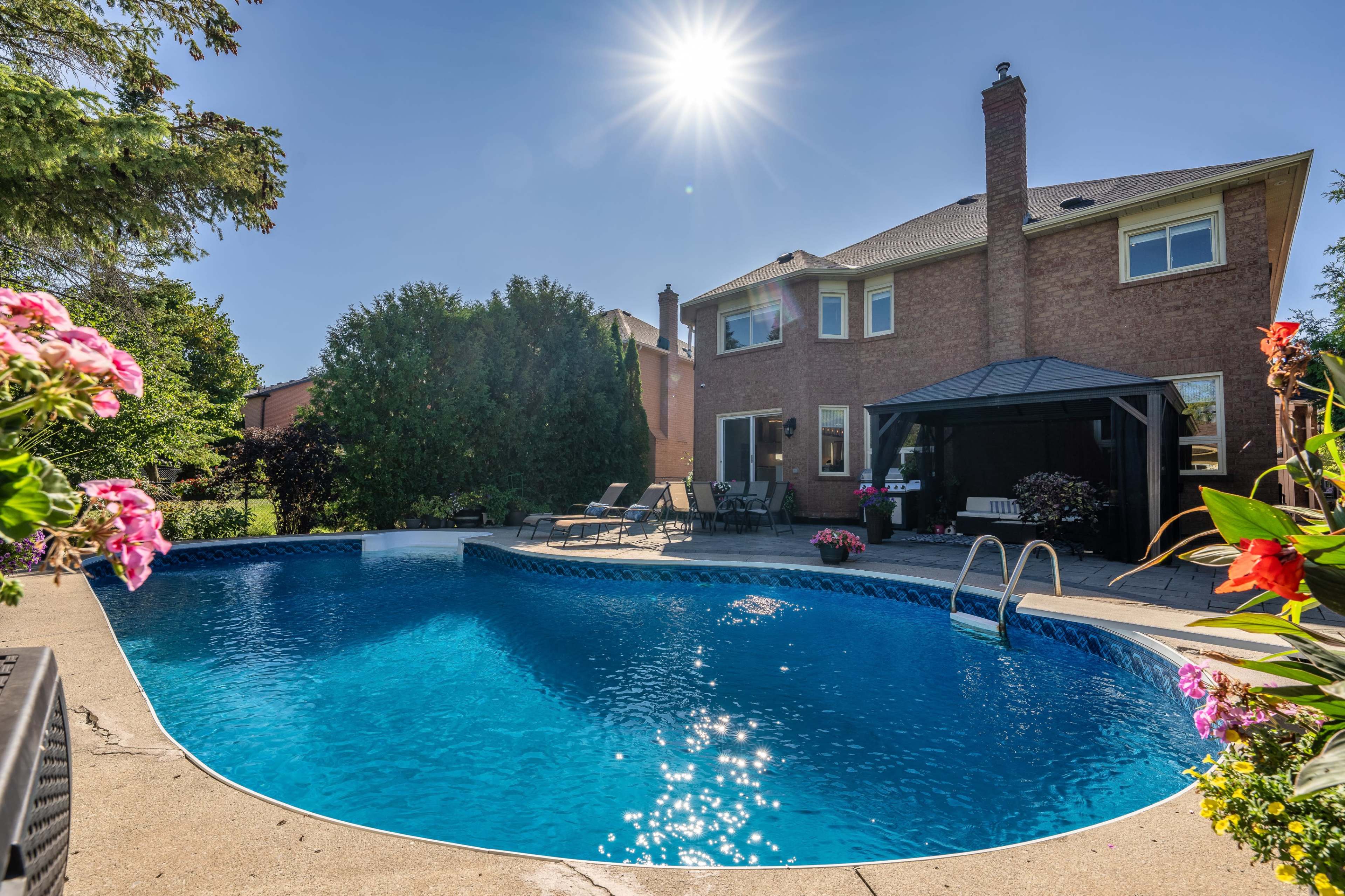 The image shows a clear blue swimming pool in a backyard with a brick house and a gazebo in the background, surrounded by greenery and flowers.