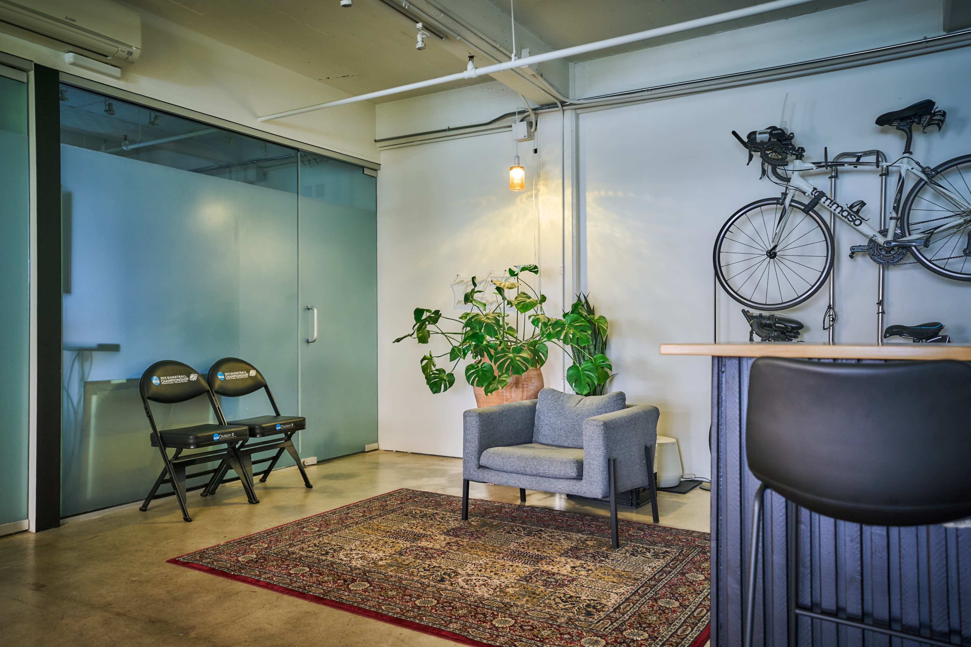 The image shows a modern office space featuring a gray armchair, a potted plant, two folding chairs, and bicycles mounted on the wall.