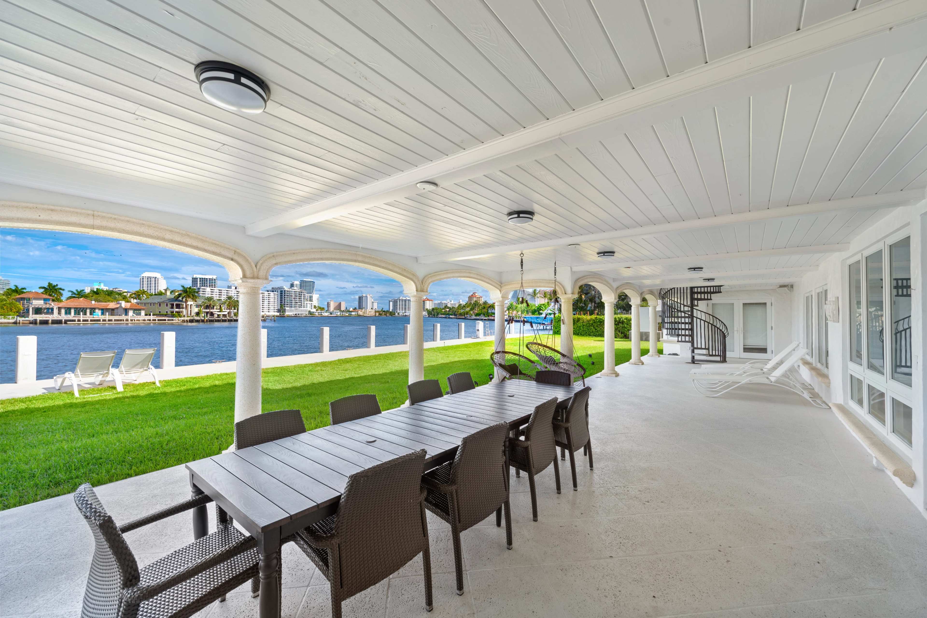 A covered patio with a long dining table and chairs overlooks a waterway and city skyline.