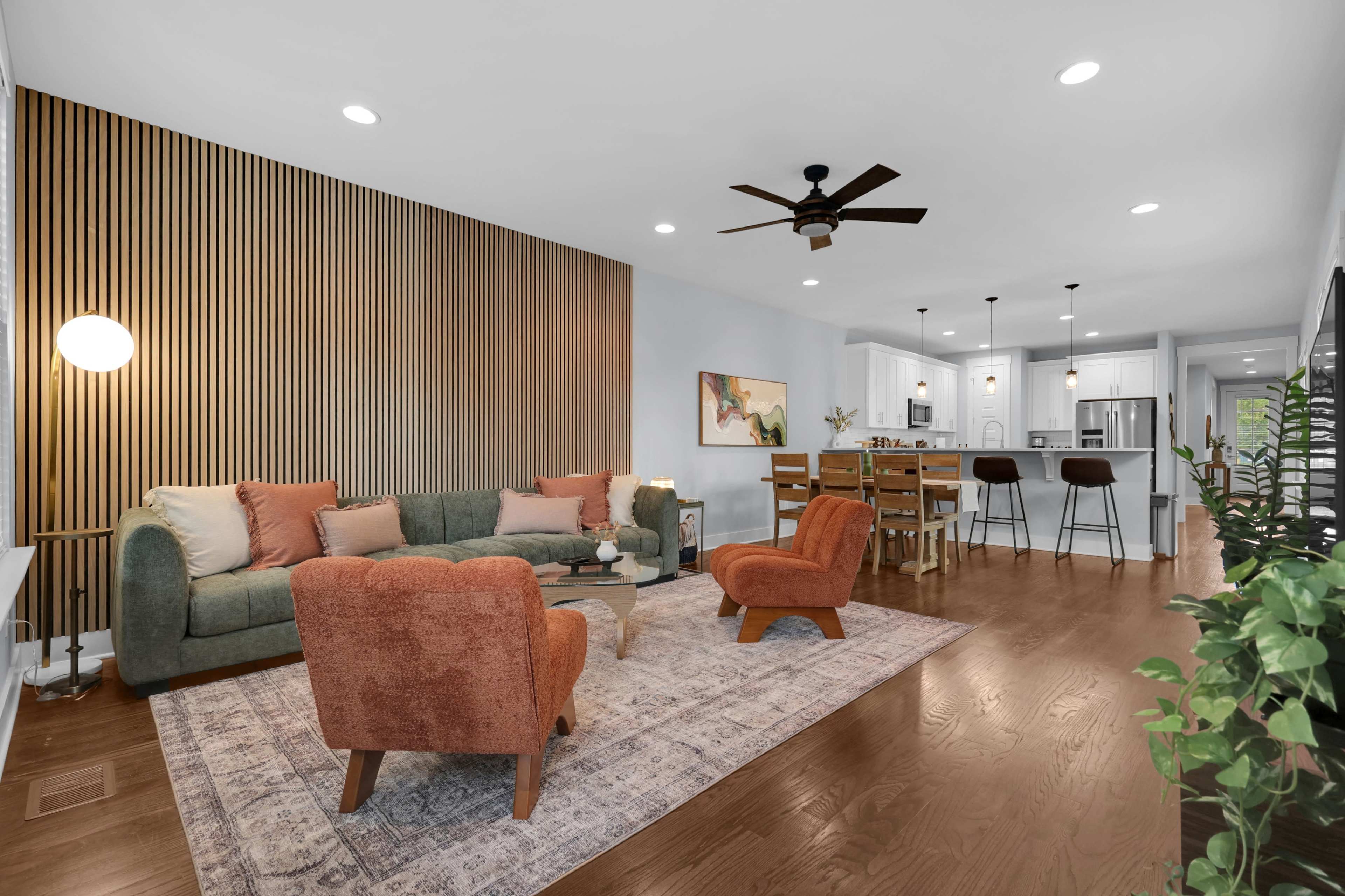 The image shows a modern living room featuring a green sofa, orange accent chairs, and a dining area with high chairs and a kitchen in the background.