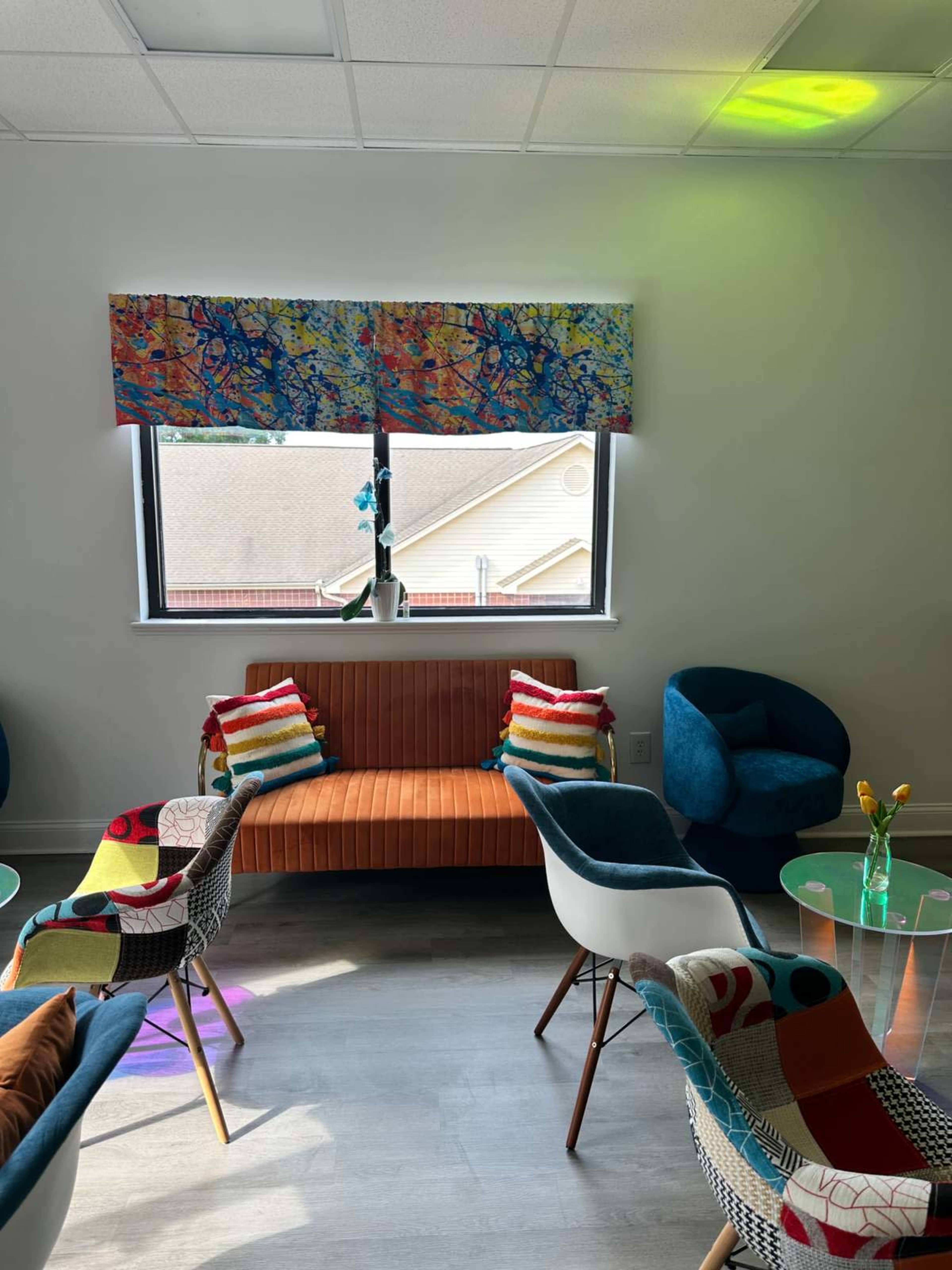 The image shows a brightly colored lounge area with patterned chairs, a striped couch, and a window with a decorative valance.