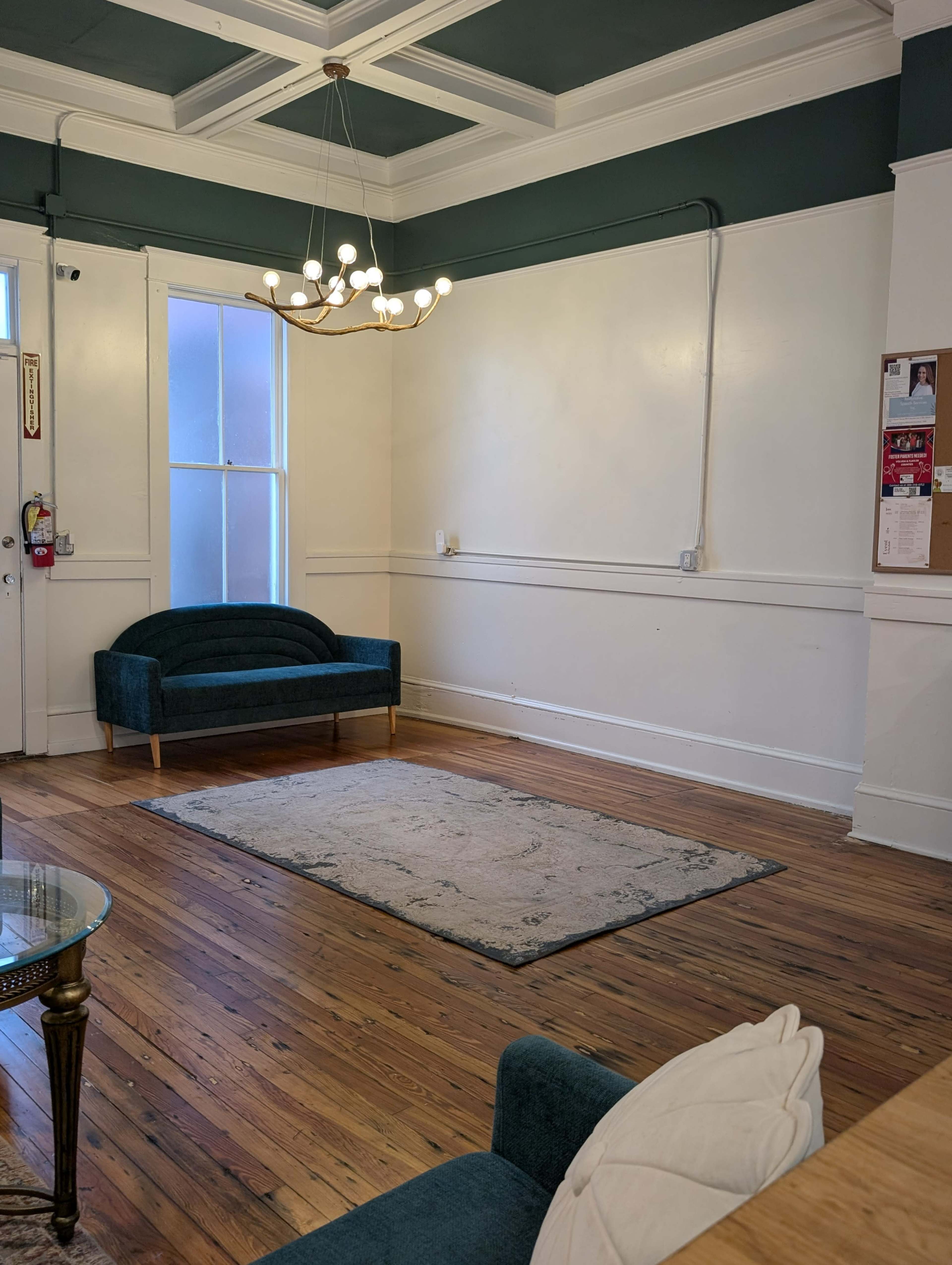 The image depicts a spacious room with wooden floors, a small area rug, a green couch, and a chandelier hanging from the ceiling.